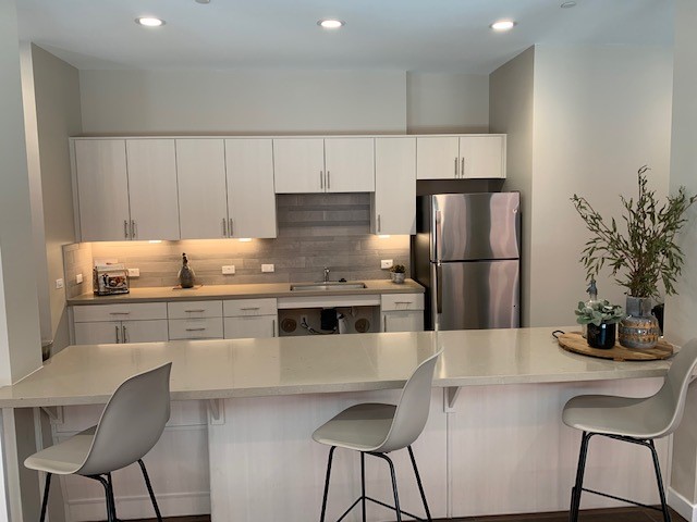 Community kitchen with 3 stool along the island.  Cabinets, a stainless steel refrigerator, and a wheelchair accessible sink in the background.