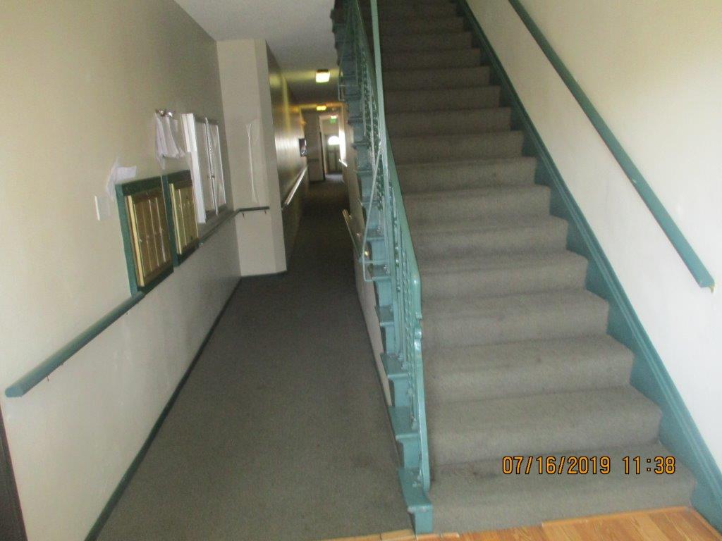 Gray carpet in well lit interior hall, on the left side wall a locked showcase, clipboard with papers, and golden mailboxes, to the right side a long gray carpeted stairs with dark aqua color handrail.