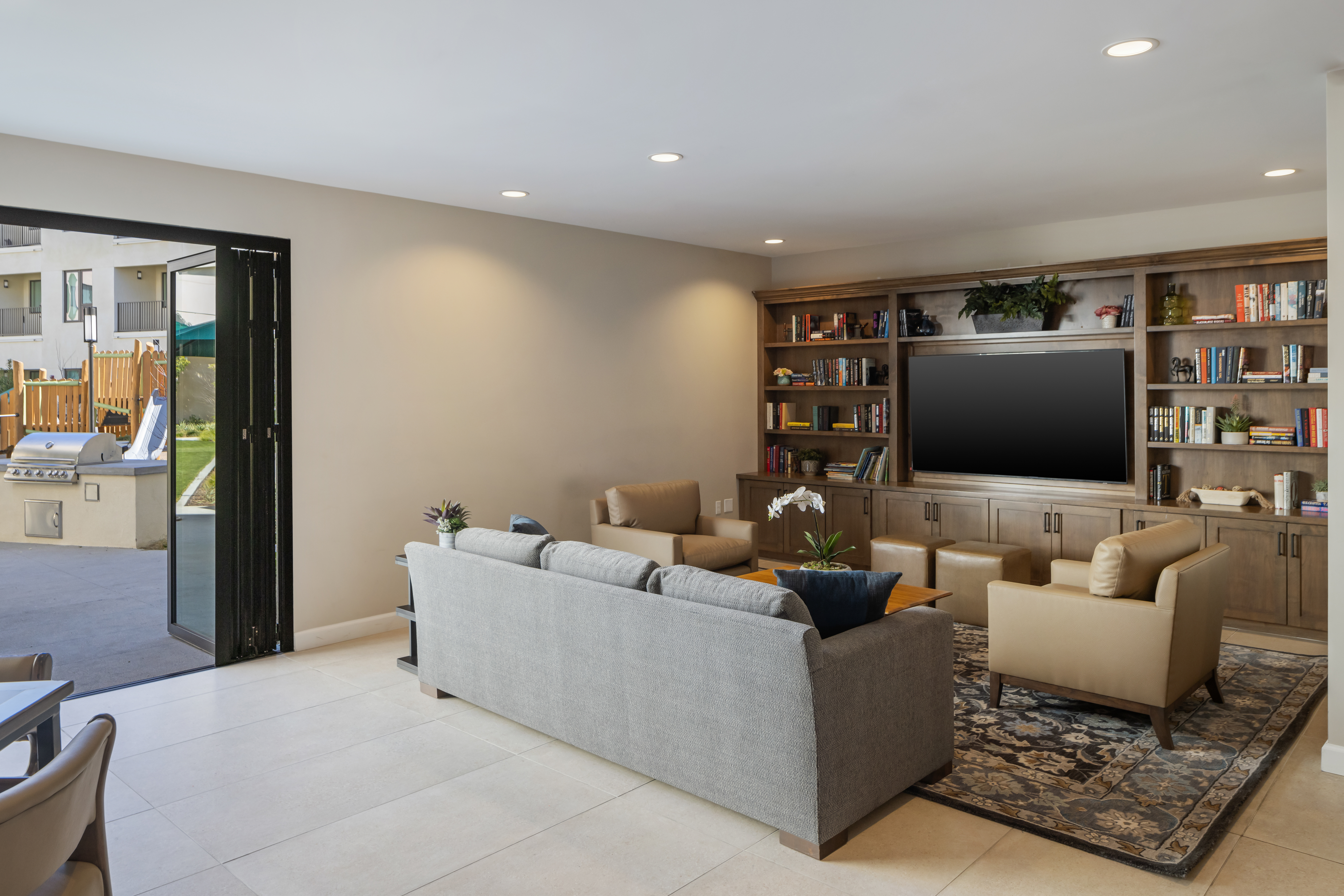 Modern community room with sofa, armchairs, large built-in shelving unit with TV, and open doors leading to an outdoor patio area.