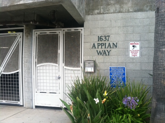 Front view of the main entrance to building 1637 Appian Way, white iron mesh doors, call box to the right side, Notice signs on the right side, plants and flowers on the right side.