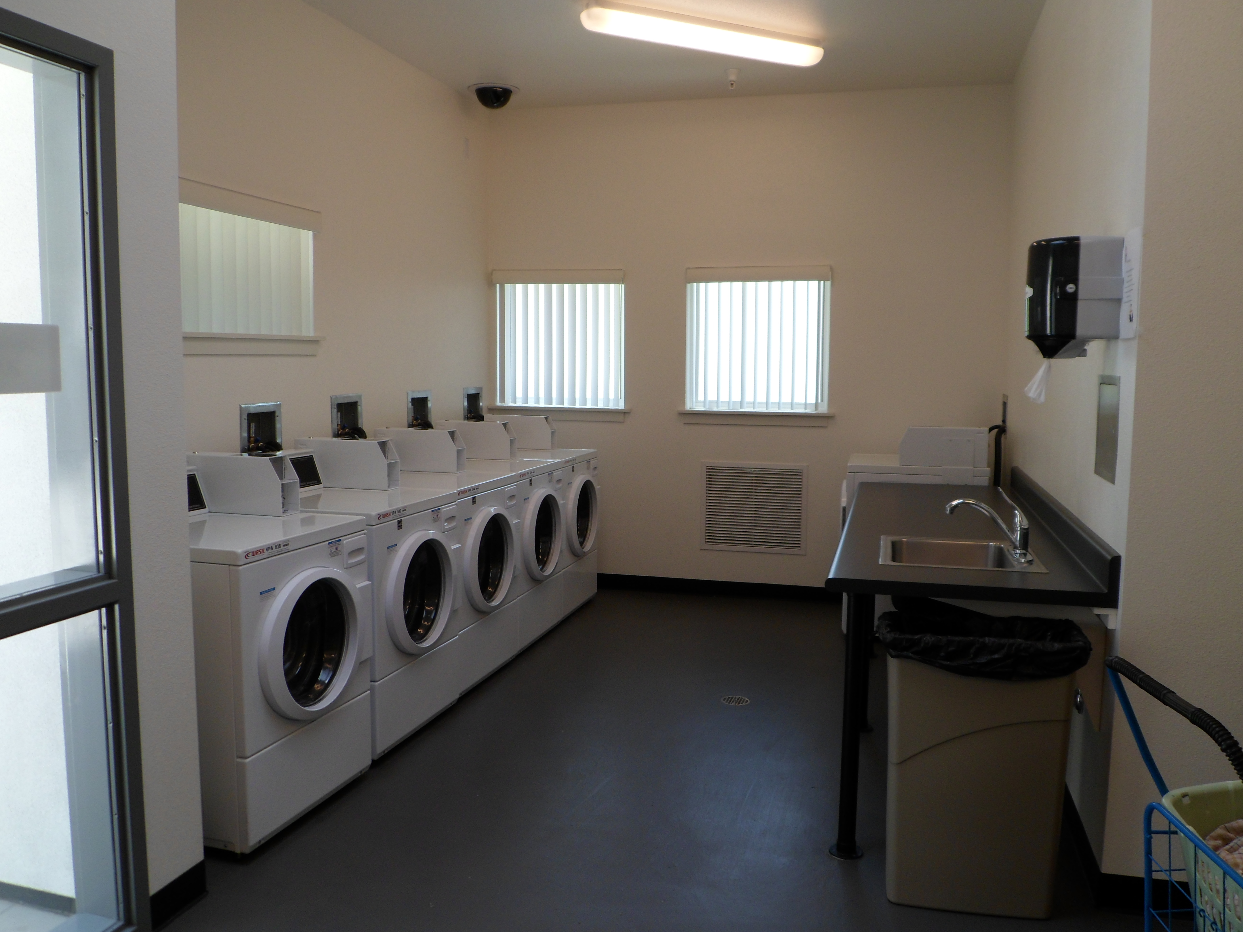 Interior view of a laundry room at Rio Vista Apartments. Five machines and a table and a sink with counter space