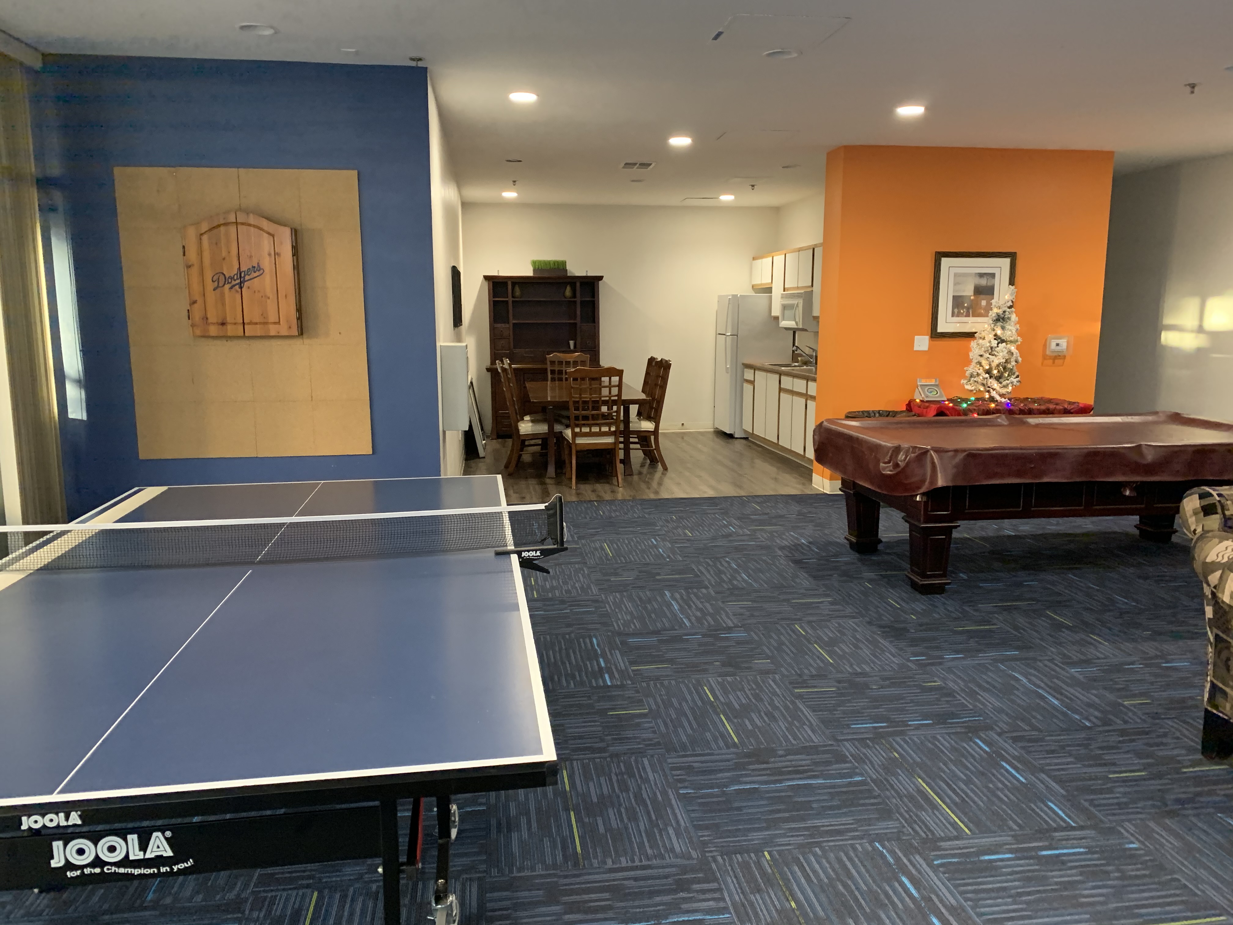 Different angle of the community room, From this view, there is a ping pong table, a dart board, a pool table and a kicthen area with a dining set table.