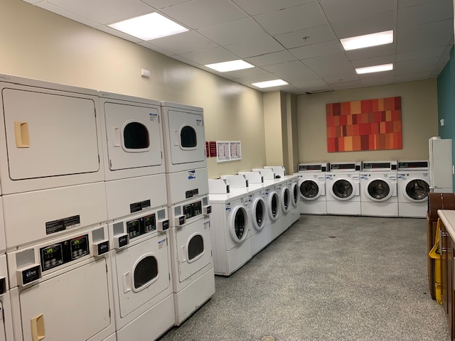 View of a big laundry room, multiple white washers and dryers, gray flooring, a colorful big picture frame on the wall, partial view of a counter with a sink.
