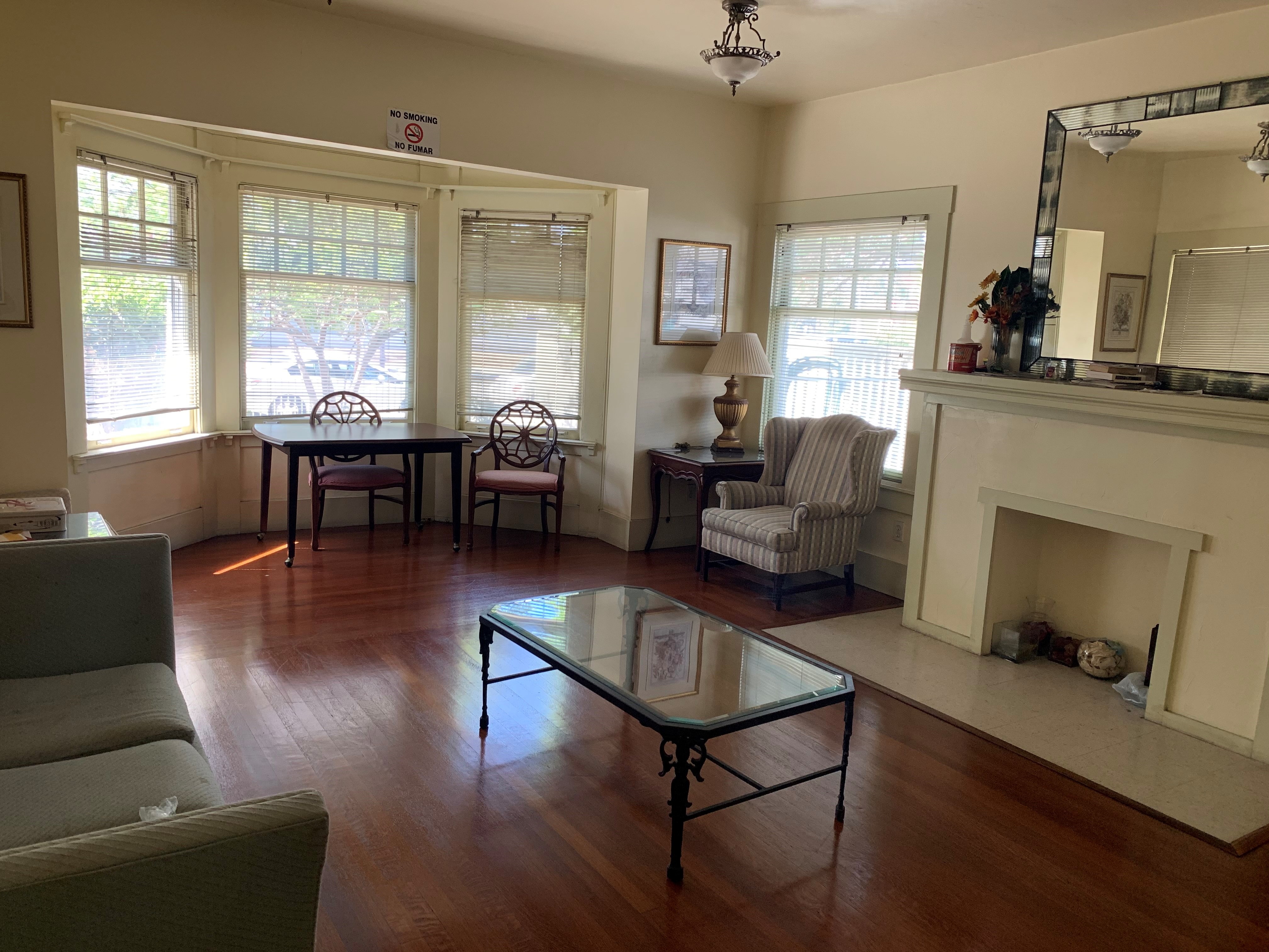 View of a lounging room with a couch, sofa chair, coffee table, a covered fireplace with plants on top, and a mirror. Floor is wooden.