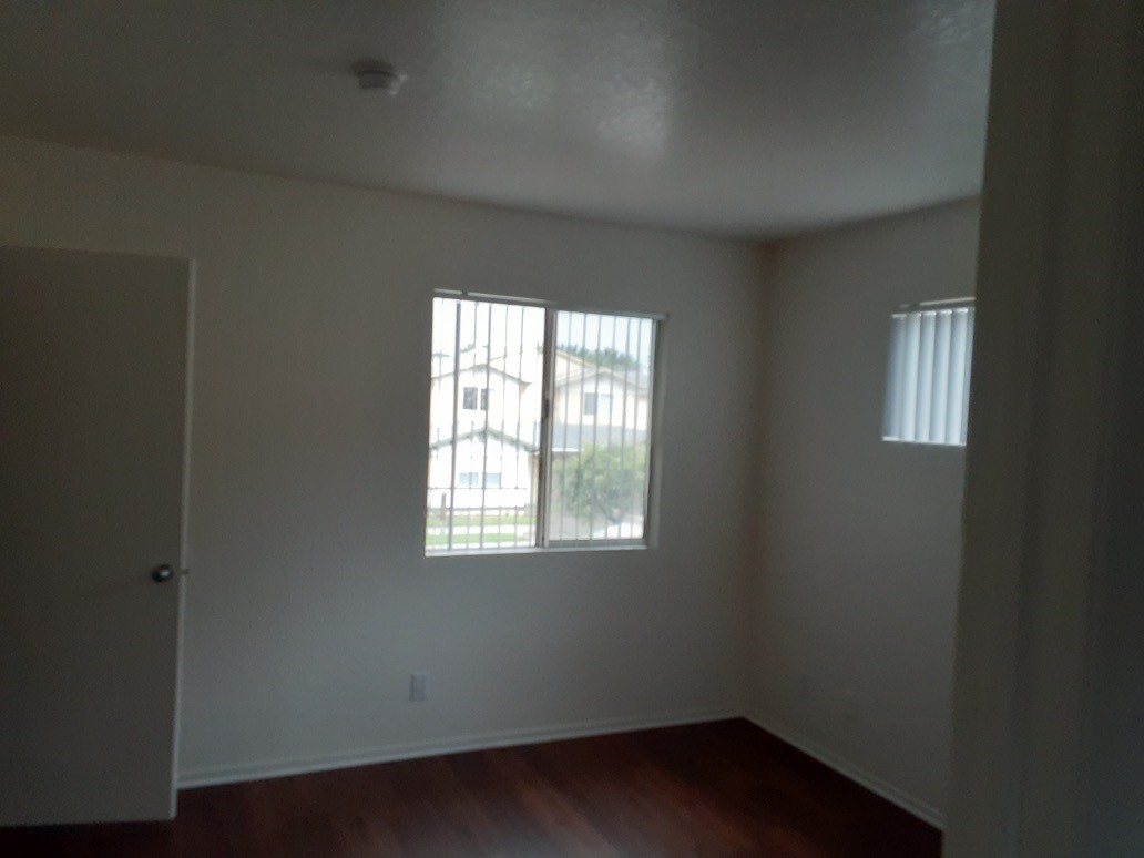 Corner view of a empty room, brown laminate floors, white walls and door, two windows with white vertical blinds.