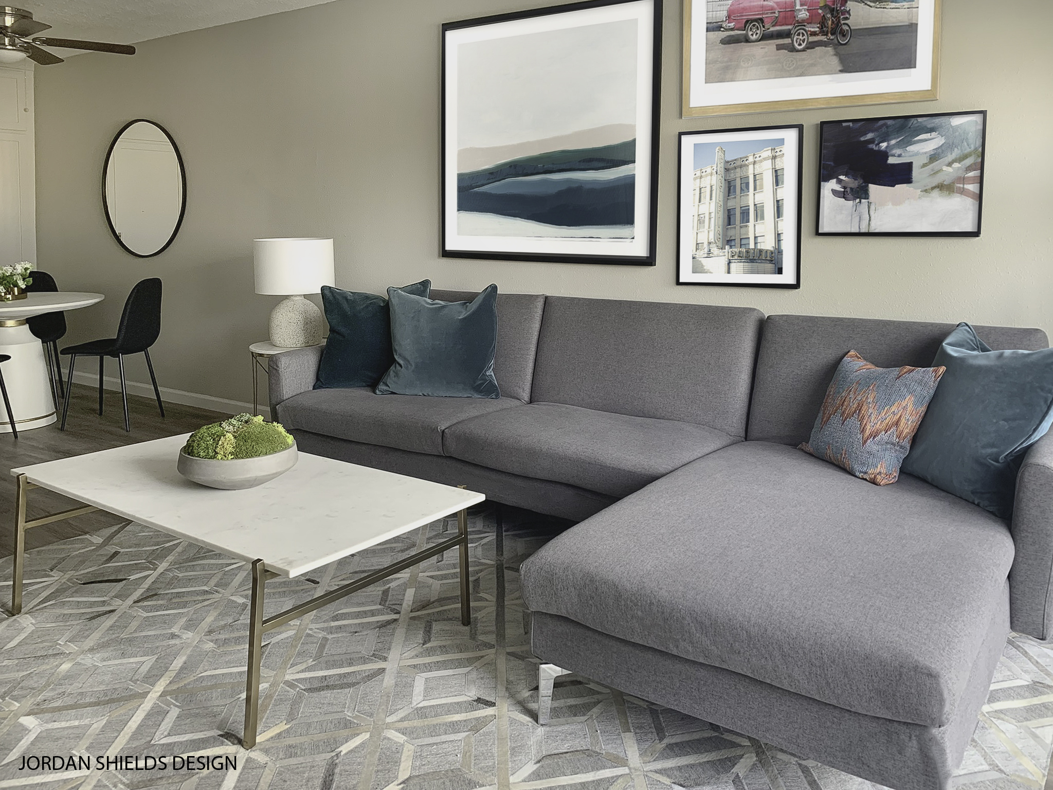 Image of room in San regis. Area has grey sectional with blue pillows over grey and white area rug. White coffee table. decroative pictures over sofa area. white round kitchen table and black chairs in dining area