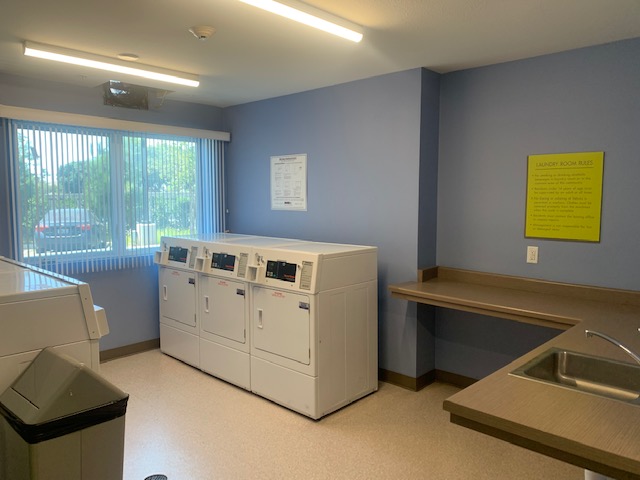 View of a laundry room, three white washers, a floding table and a sink, beige trash can, a window with white vertical blinds.