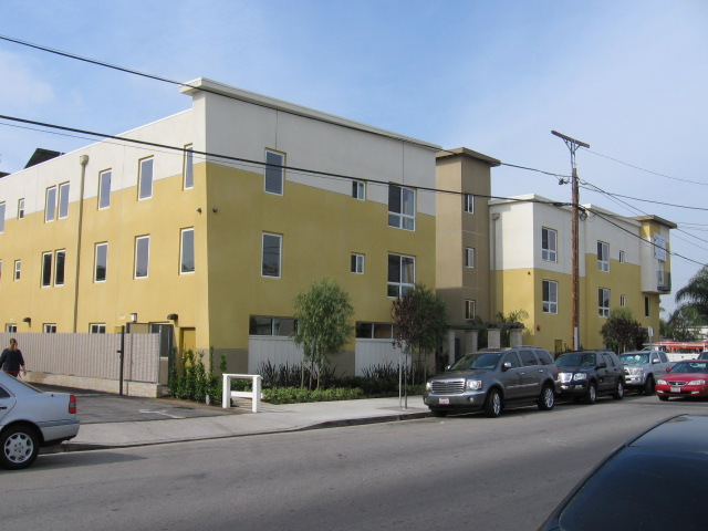 Side view of 3 floor apartment building, green shrubs and trees along building and street parking in front of building