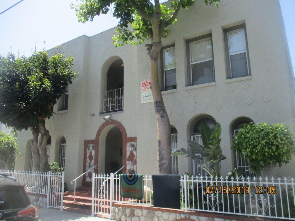 Front view of building, multiple windows, bottom windows gated, one balcony in the top middle of the building, trees right in front, white small steel fence with open entrance, closed to the public and building name and phone numbers signs, wide five step