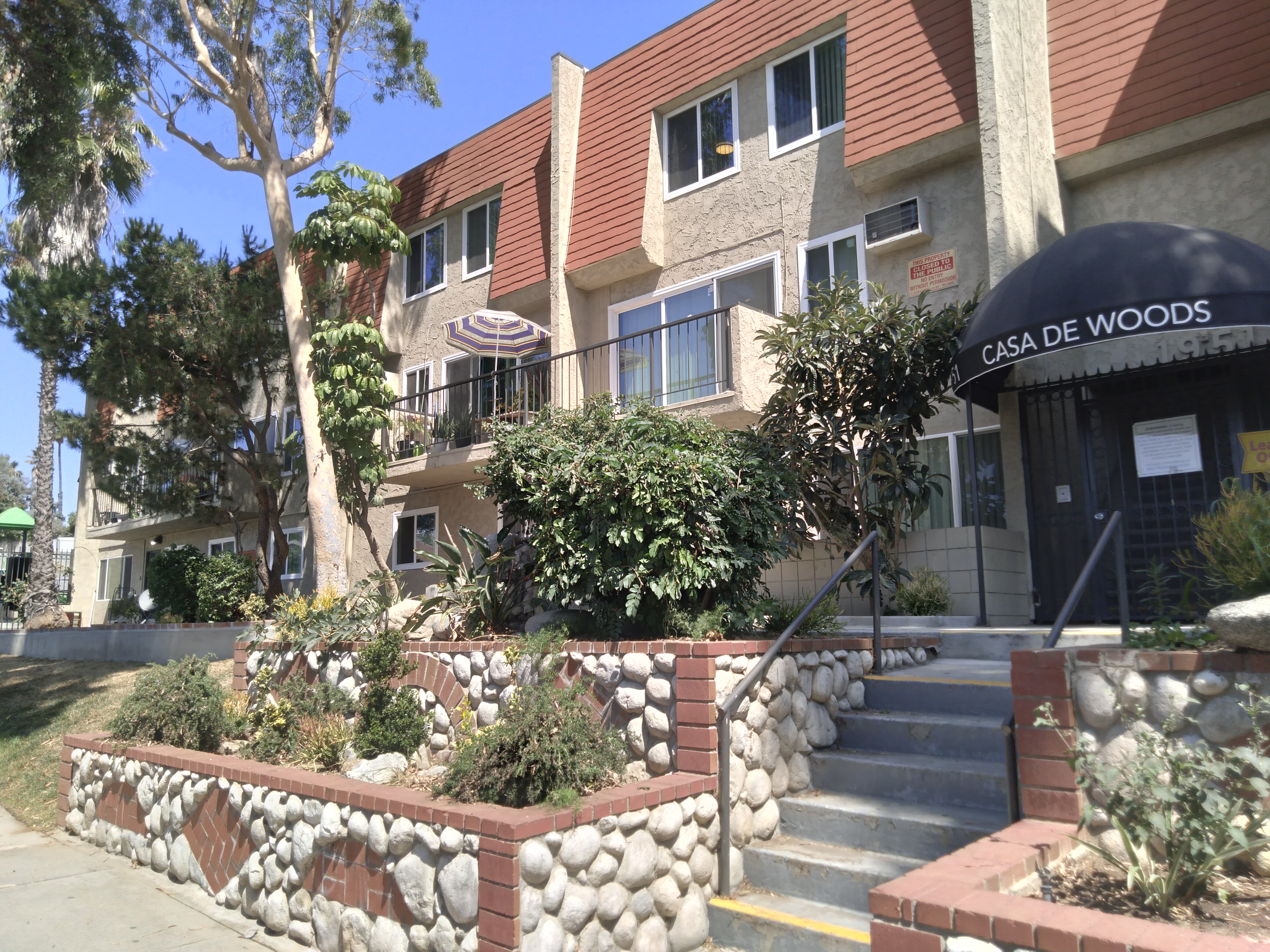 3 Story beige and red orange brick color, six stairs up in the front entrance with black iron handrails, landscaping in the front of the building, white stones designs pillars