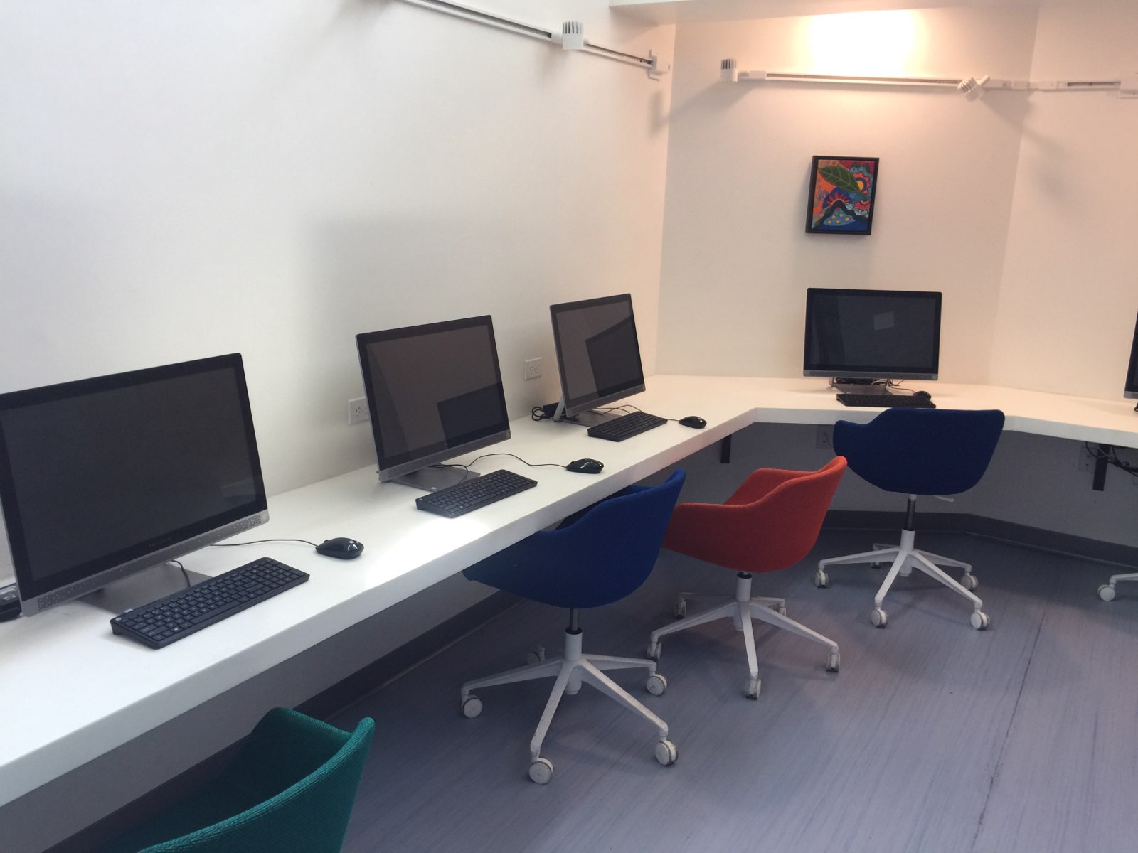 Computer room with multiple desktops, different colored rolling computer chairs, a small painting on the wall.