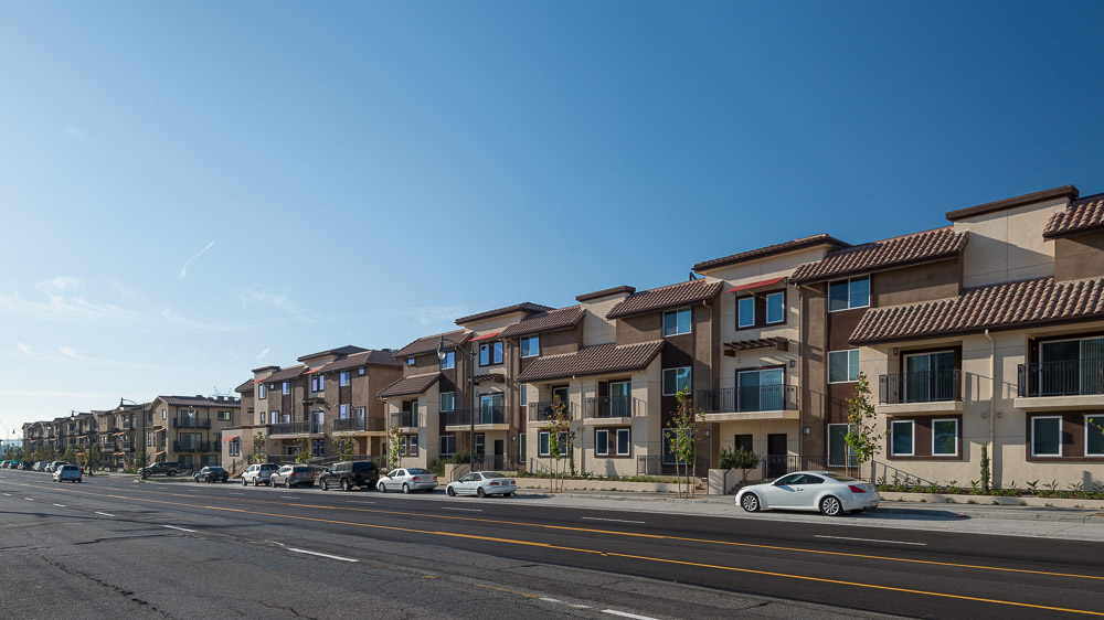 Exterior street view from San Fernando Road of Rio Vista Apartments. 3 story Earth tone building with cars parked along the street.