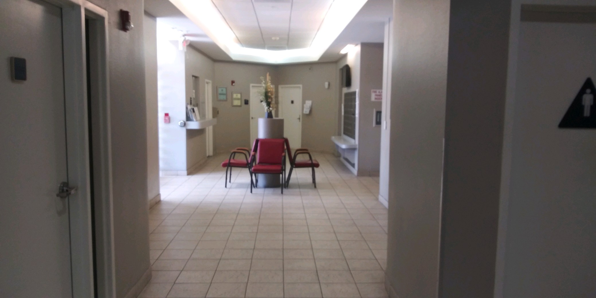 Side view of a lobby area. Room contains red chairs that are wrapped around a centerpiece with a plant that sits on top of it. There is an office window on one side of the room and three doors adjacent to it. There is a restroom entrance pictured.