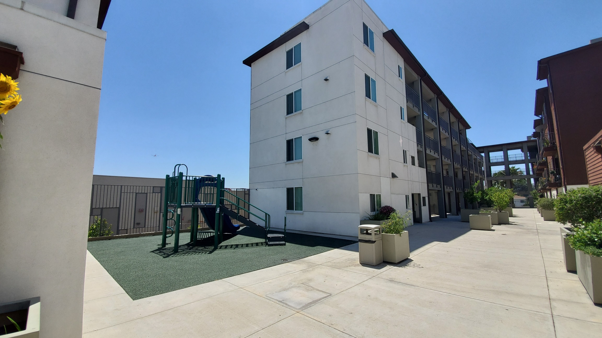 Different anlgle of a playground that is inside a complex. Playground is in an open space. It consists of a slide, stairs, and a climbing ladder. The ground is padded. There are adjacent units on both sides of the playground, and a variety of plants in re