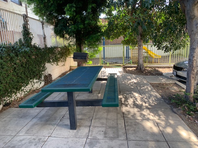 View of a picnic area within the building near the gate and parking lot. The ground is concrete with a park table,  benches, and  bar-b-que pit.