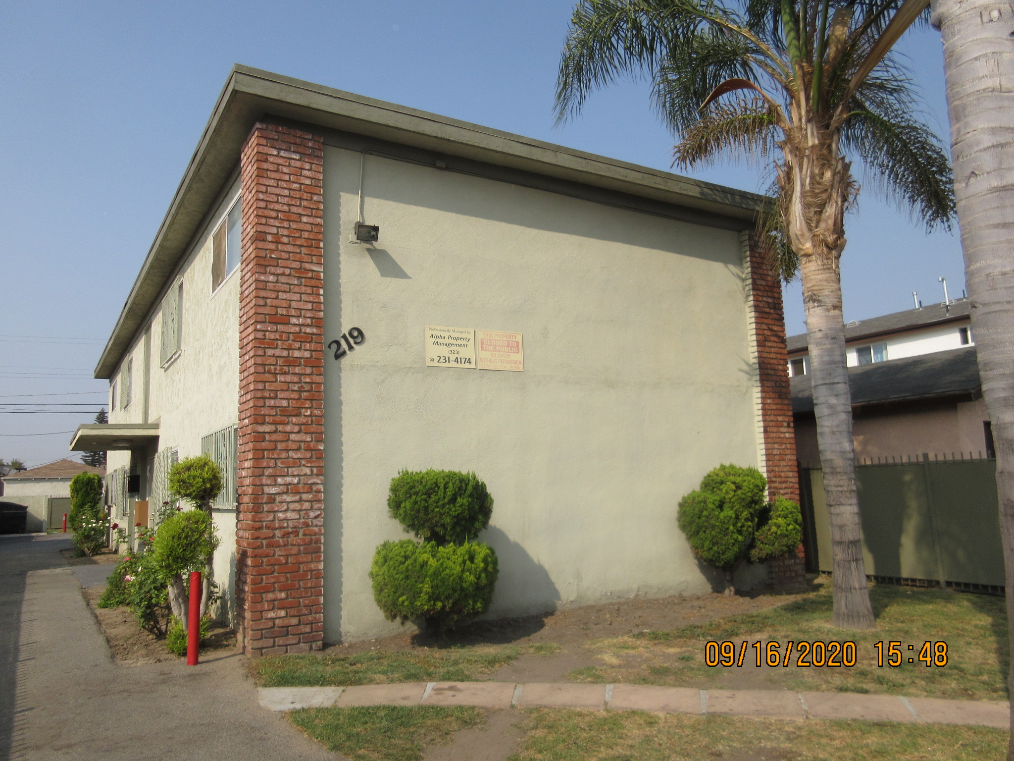 Right side view of a light green two story building, building number and management signs on the wall, palm trees and bushes.