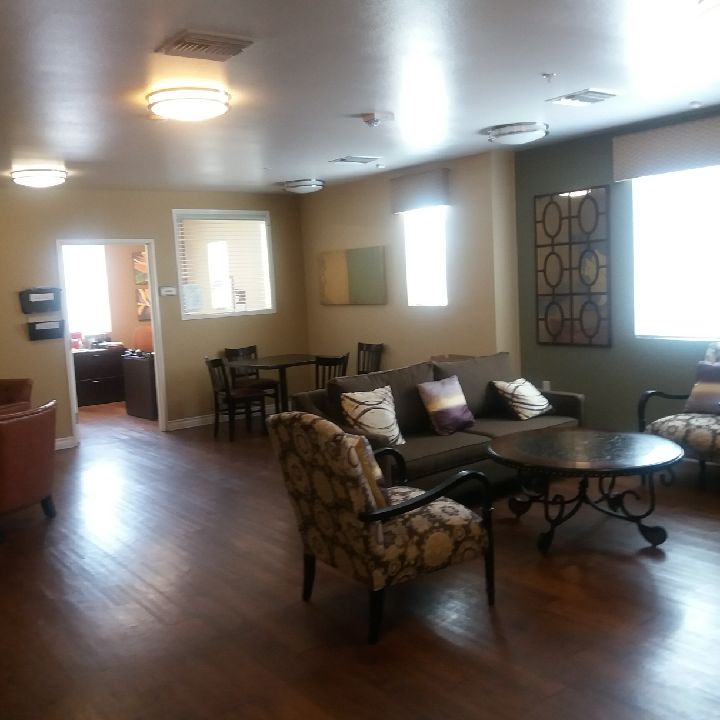 Community room with a small dining set table, two sofa chairs and a coffee table, wood flooring, abstract art on the wall, yellow/earthtone/muted-blue color scheme, view of the leasing office.