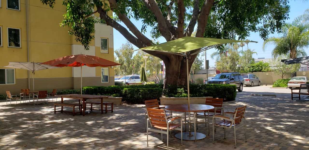 Rayen Apartments courtyard. Within courtyard there are several patio table and chairs that have umbrellas for added shade. A large tree incircled by small bushes located near parking area of courtyard.