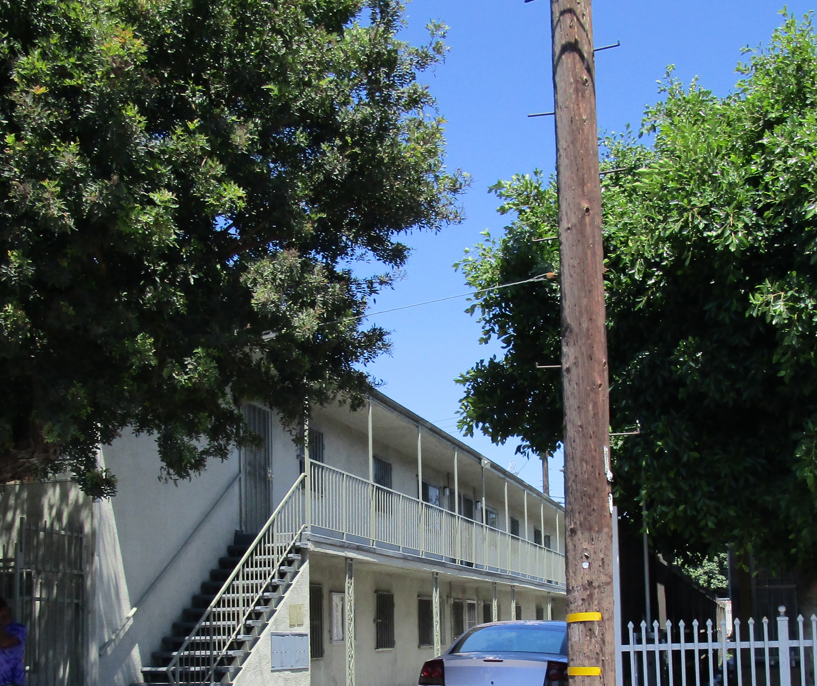 Side view of a white two story building open, no walls, a long staircase with hand rails, a gate all along the units, Open parking entrance, parked car, a big tall tree on the right side and a light pole tree on the left side, gray mailboxes.