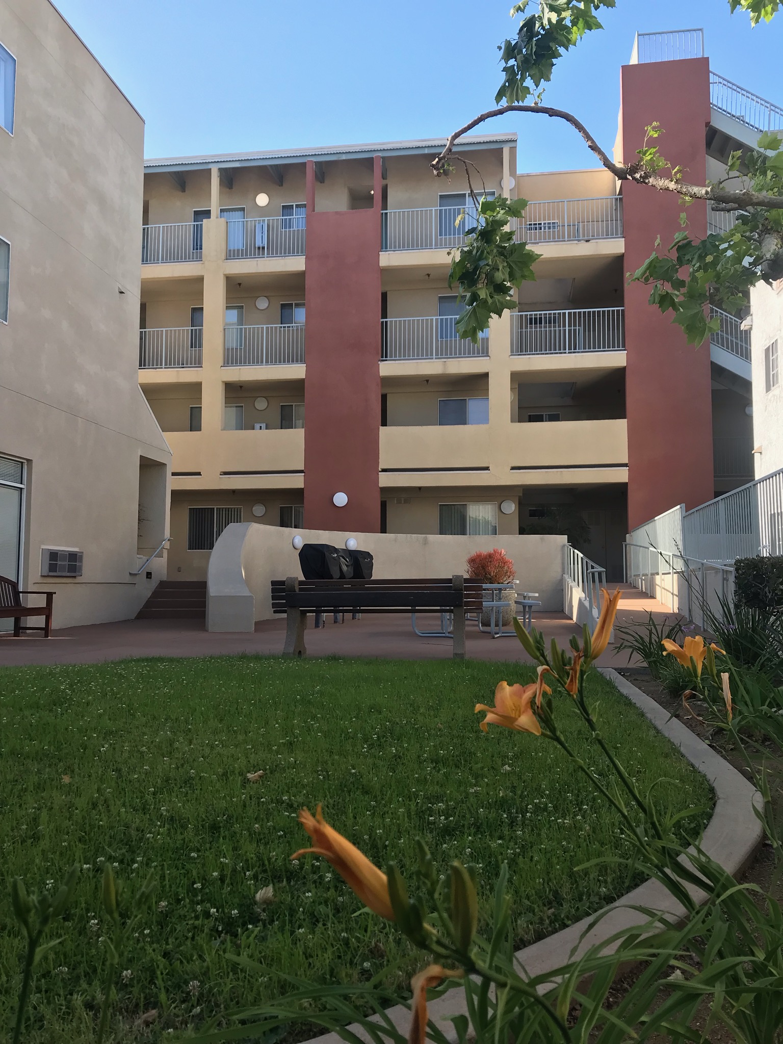 Four story building with an outside community lounge area.
Steps and accessibility ramp that leads down to bbq area 
with bench seating and patio table. Small grass Next to 
grass area is a flower bed along the fence