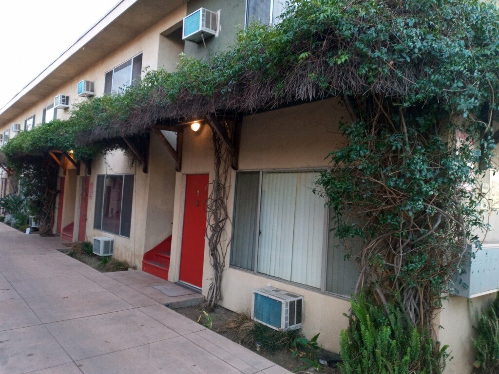 Front view of building. Red doors for units with stairs between them. Awning with greenery grown throughout.
