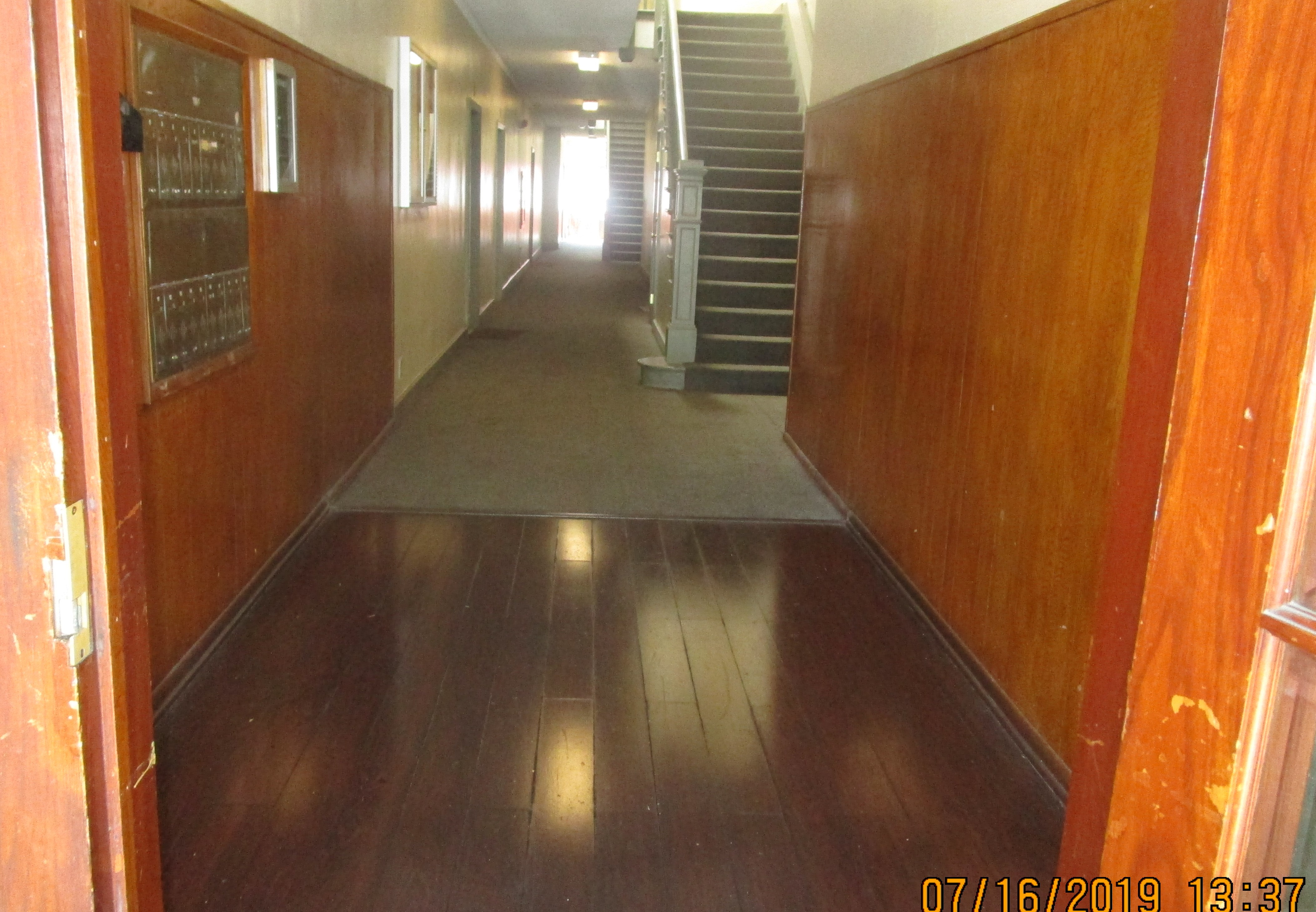 View of long hallway, mailboxes on the left side against the wall, brown wood laminate by the entrance and brown carpet on the hallway, brown carpet stairway with handrails.