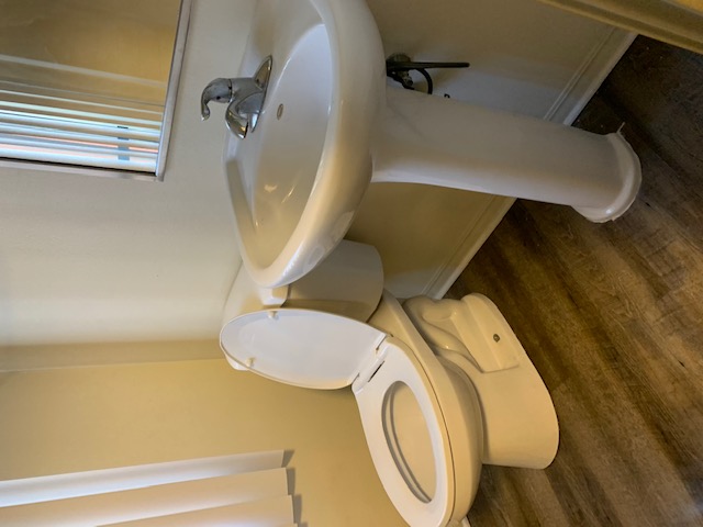 Photo of a restroom, showing mirror on the wall, sink below it and a toilet with the toilet seat up.