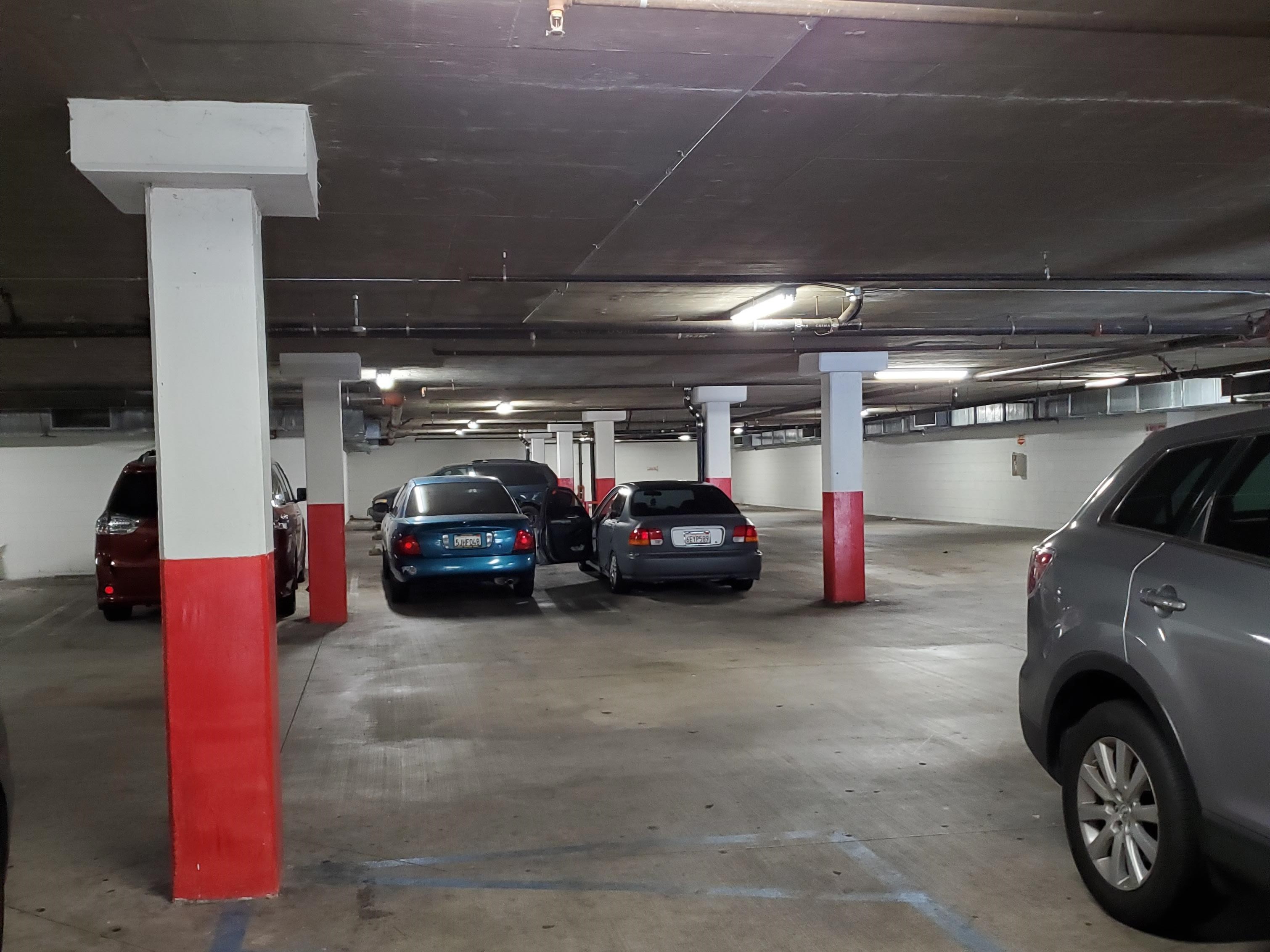 Underground garage with cement floor, four parked cars, one more car with driver side door open, wide tall pillars white top and red bottom color.