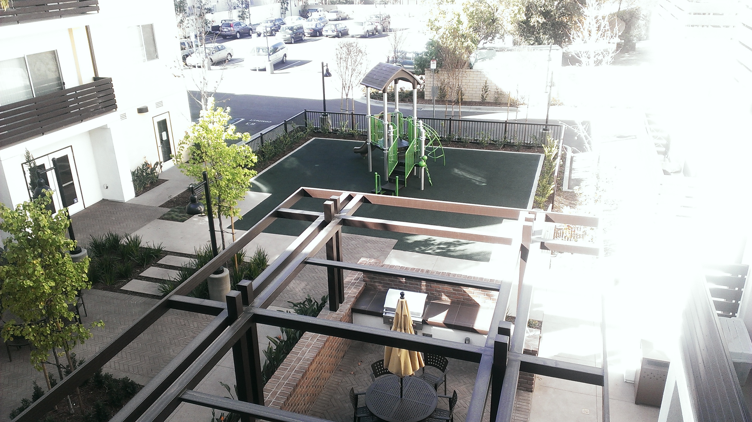 Top view of a courtyard with a small playground and a grill with a table. There is multiple plants around the area. Courtyard is adjacent to an outdoor parking lot.