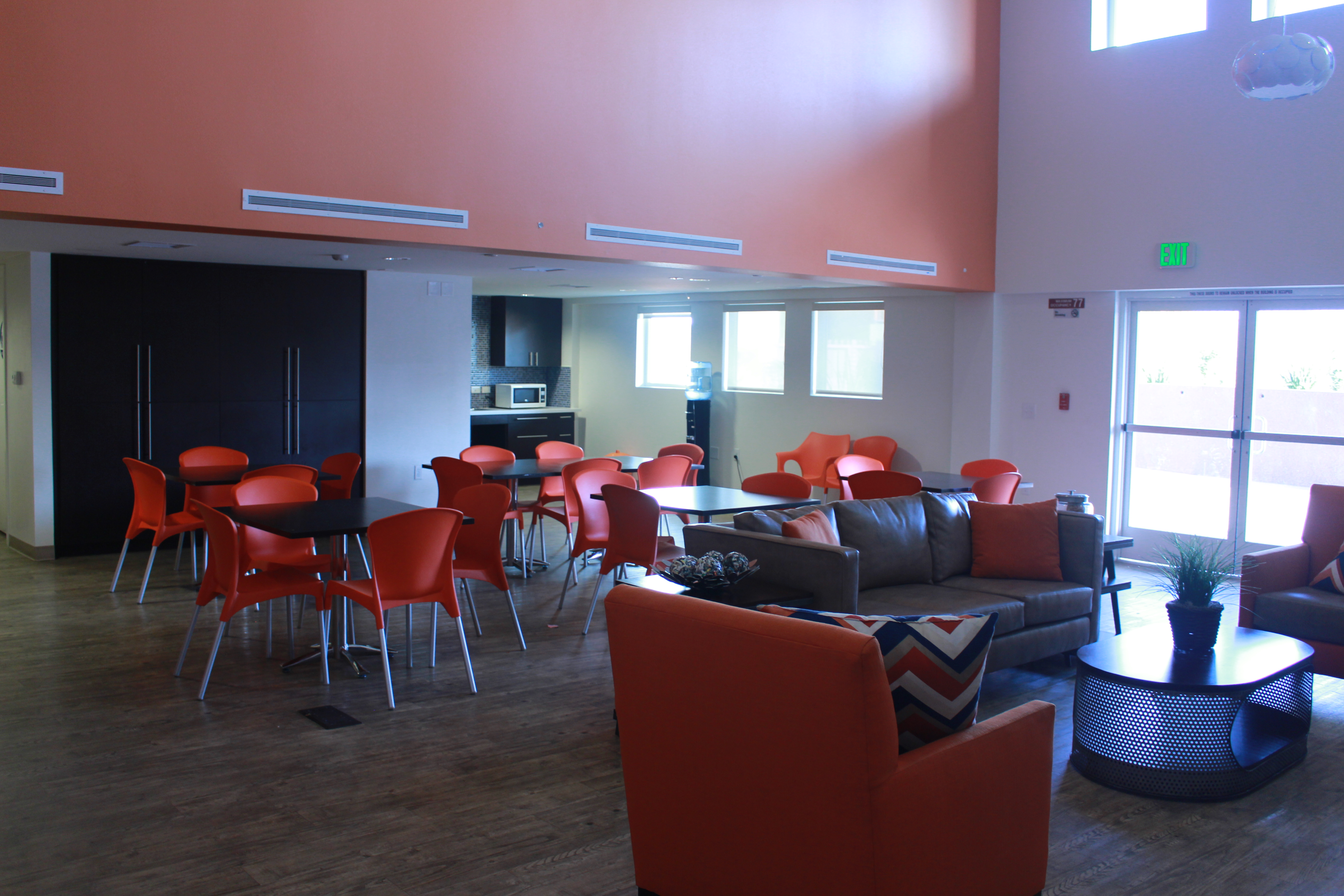 Community room, five brown square tables with four plastic orange chairs, a long brown sofa and two orange sofa chairs, brown oval coffee table in the middle with a plant base on top, multiple decorative pillows on the sofas, a counter with a microwave to