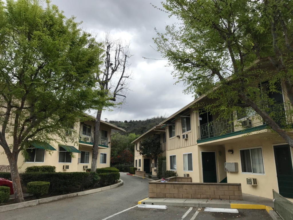 Image of identical 2 story buildings, some units with balconies and a/c units, grass, trees, plants, and parking along buildings