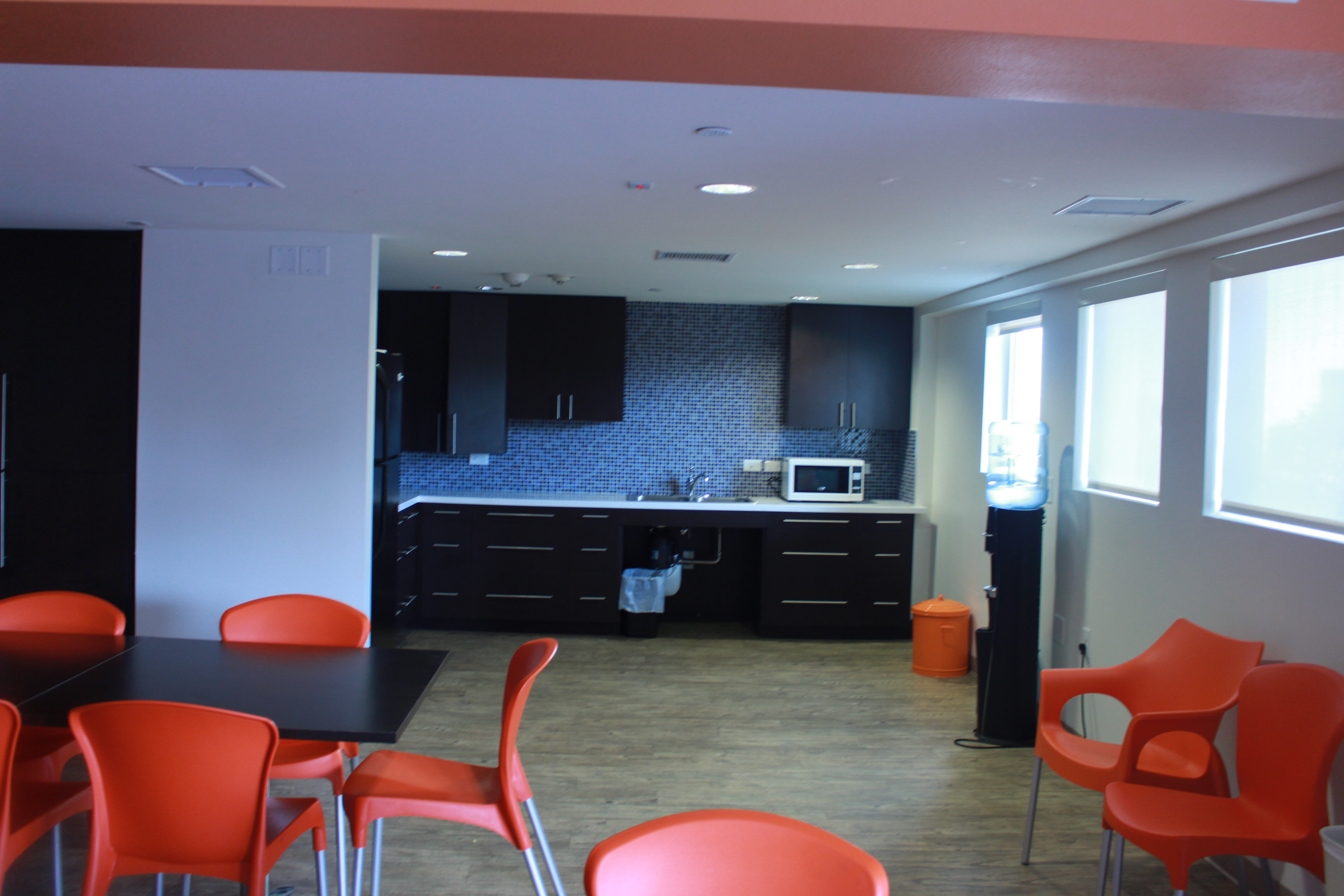 A second kitchen view, dark brown cabinets, a sink, white microwave, a trash can under the sink, black water dispenser, an small orange dispenser with lid, many oranges chairs and part of a table is visible.