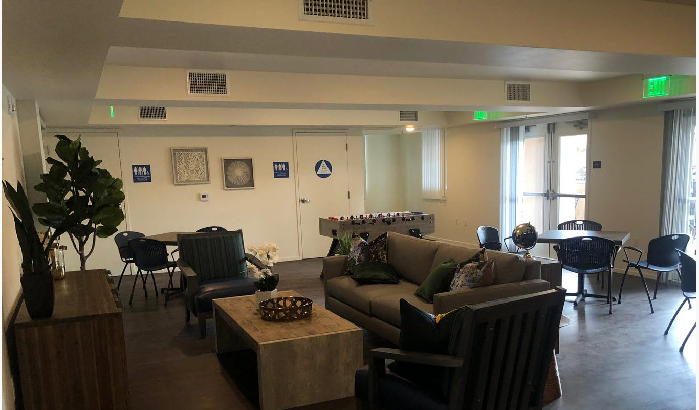 Community Room with couch and chair, two tables with four chairs at each table. One fools-ball game and two restrooms.