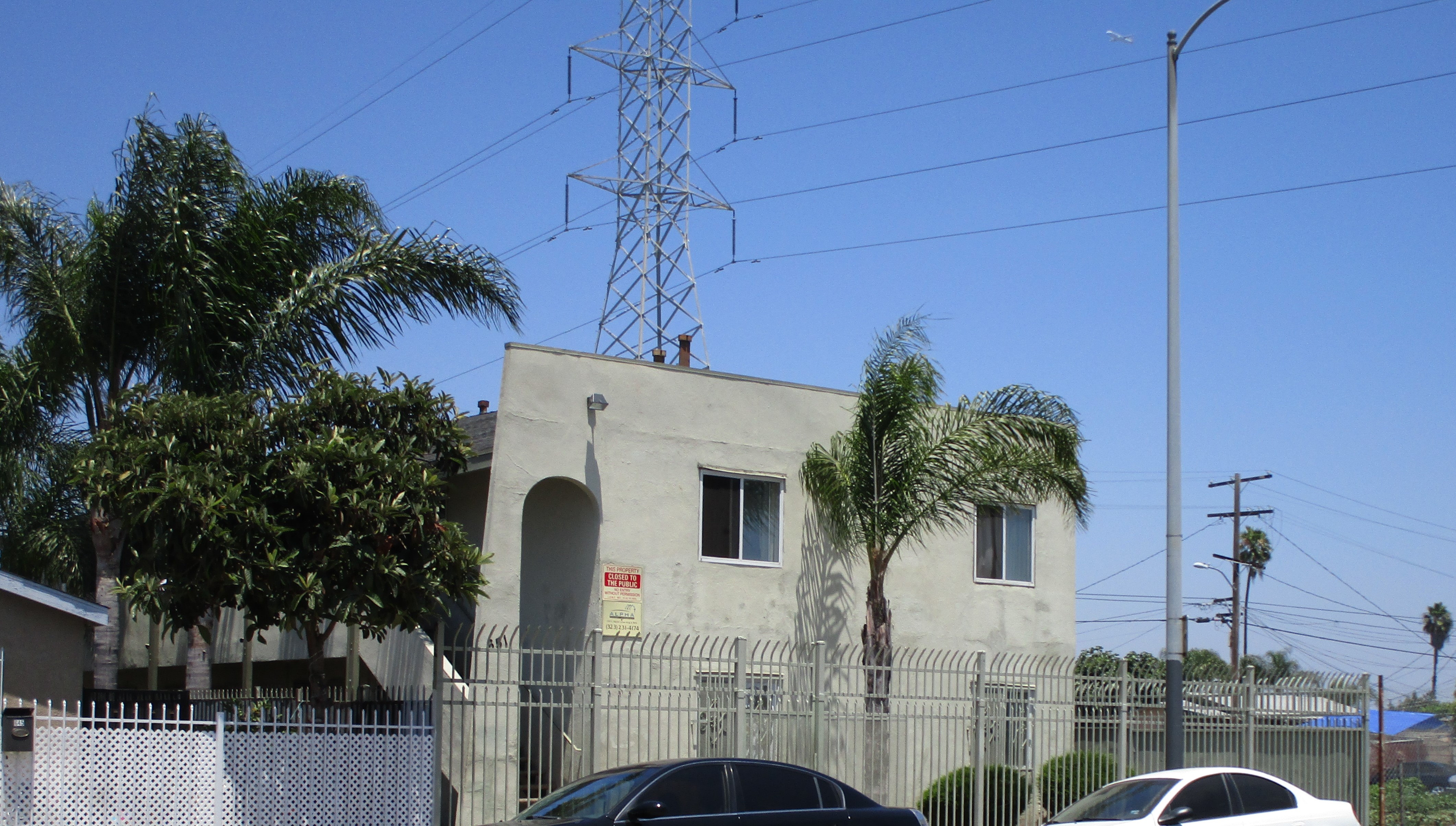 View of a two story gated building, four windows, palm tree and bushes in front, parked cars.