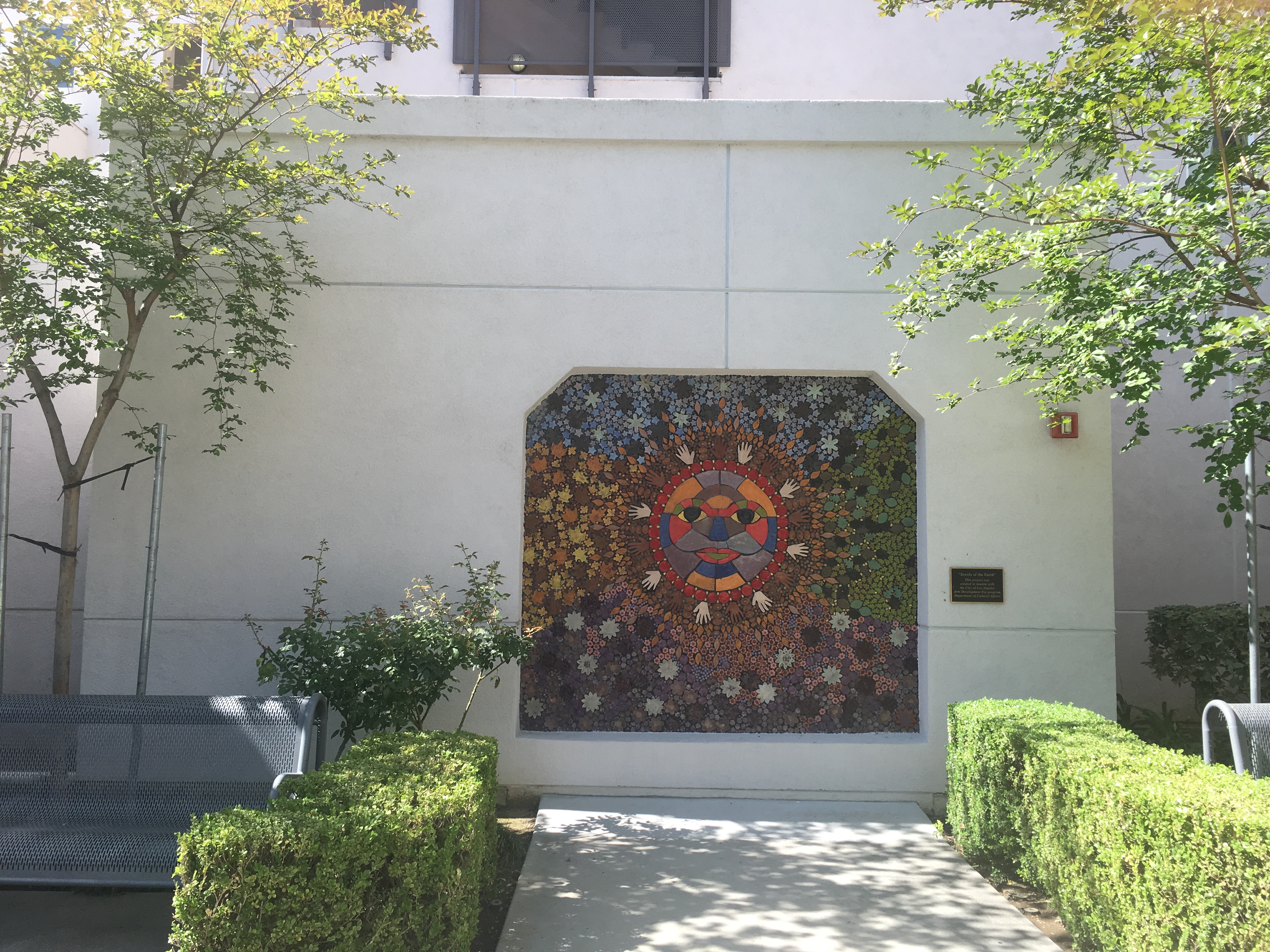 Front view of the court yard, a beautiful colorful mosaic art on the wall, well trimmed bushes on each side, gray iron mesh benches on each side.