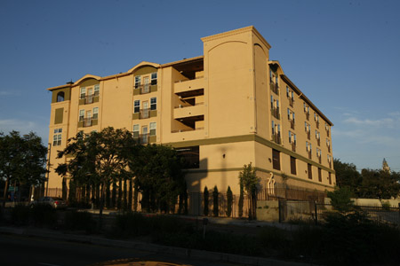 Side view of a four story mustard yellow building, multiple windows some with balconies, trees around the gated building.