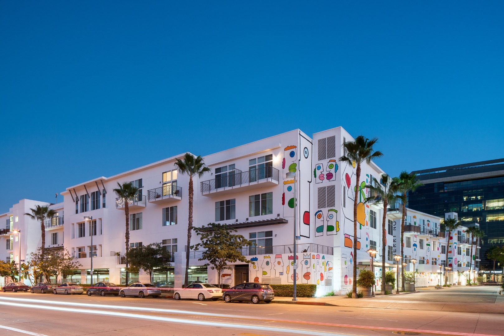 Street view of Lofts at NoHo Commons, 3 story white building with colorful abstract face murals on one side of the building and palm trees surrounding builing