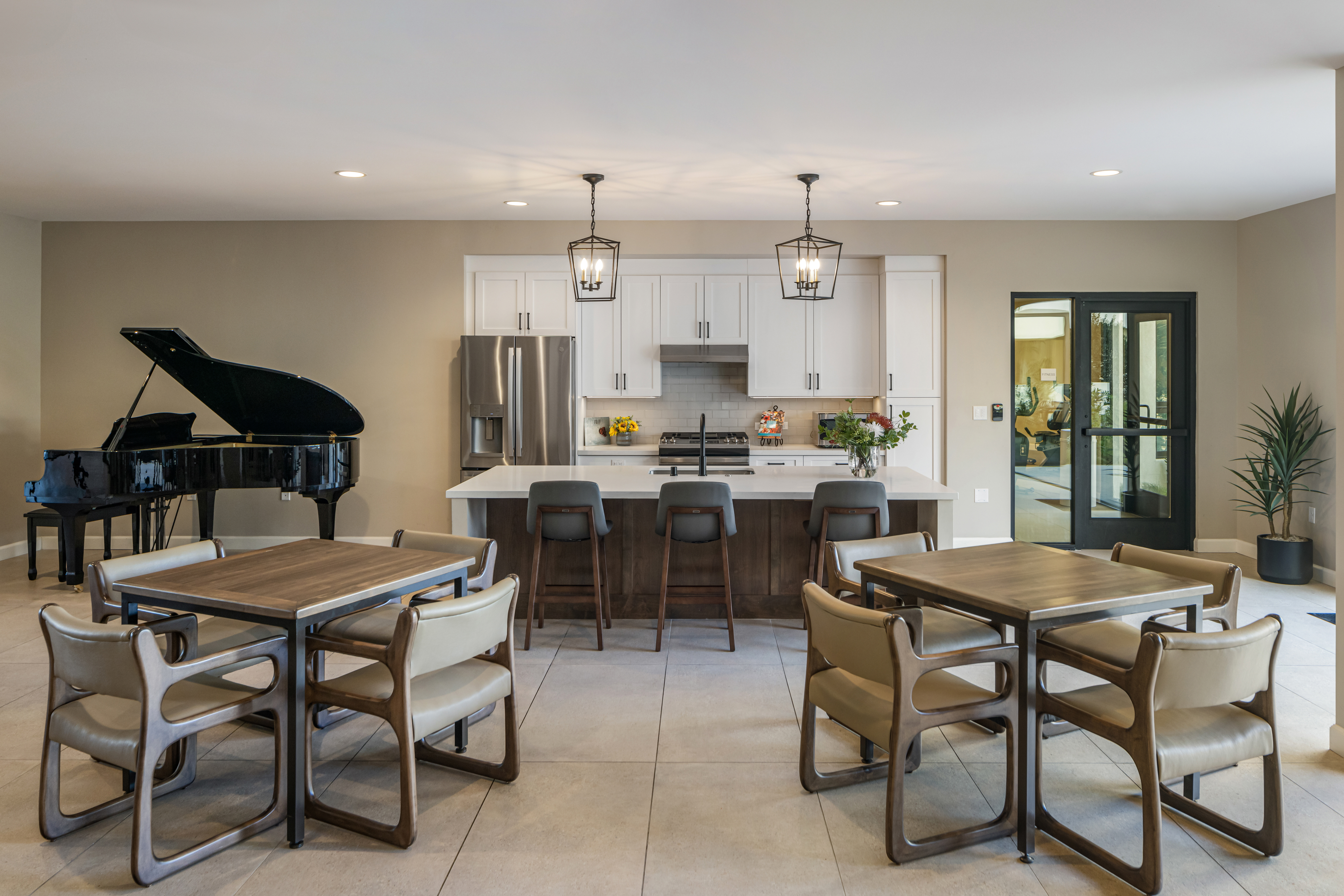 Modern communal kitchen and dining area in affordable housing, featuring a large island, four bar stools, two square tables with chairs, a black grand piano, and potted plants.