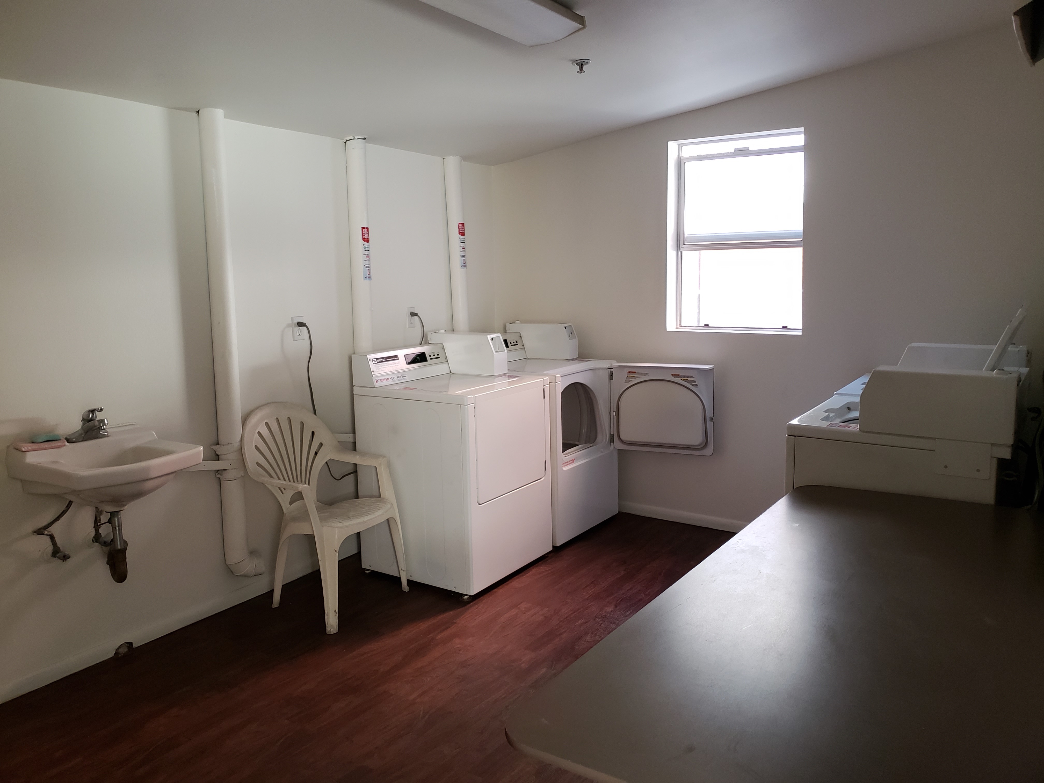 Inside view of a laundry room. Room consists of a sink, 4 machines, a table and a chair. There is also a small window.
