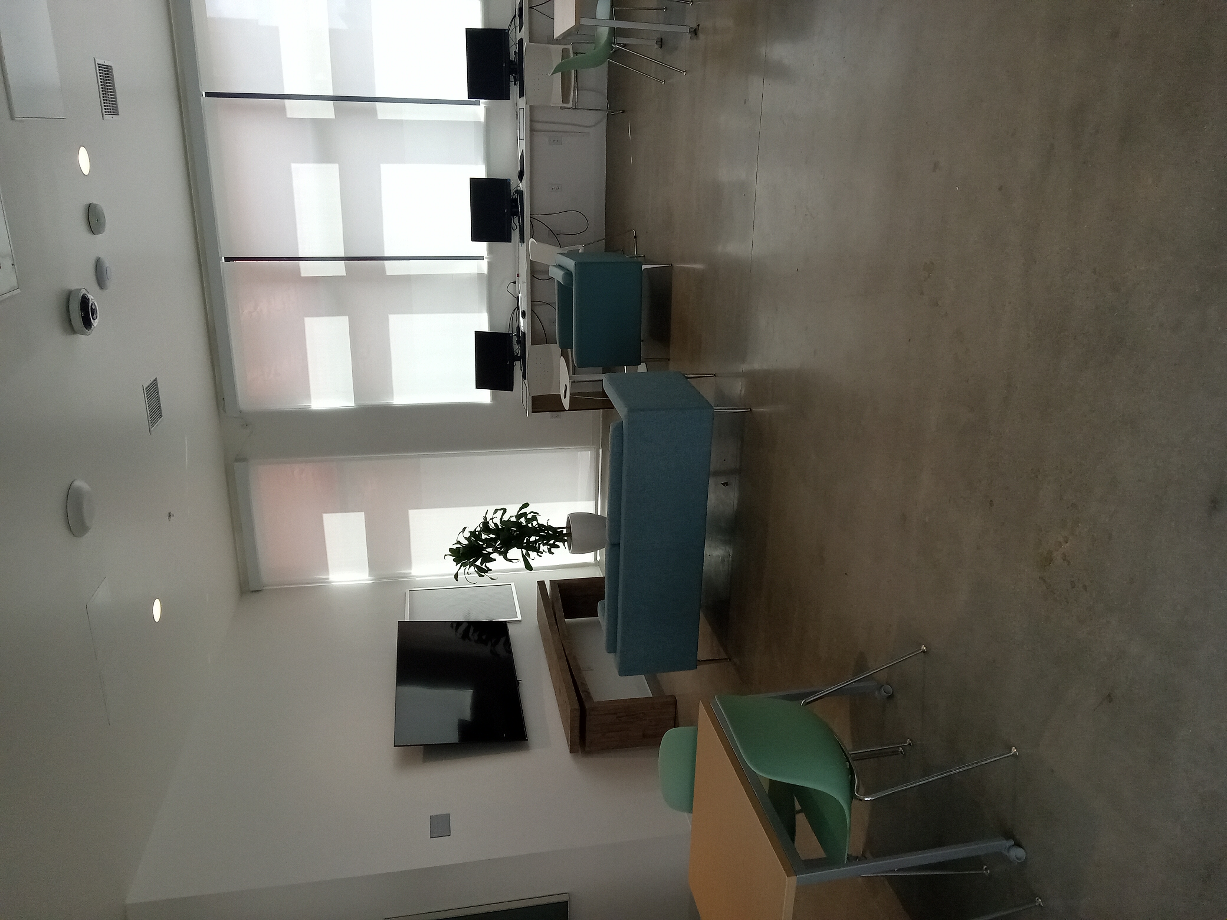 Community room: Large flatscreen tv, teal-blue couches, mint-green stackable chairs at tables, white walls, concrete floor, plenty daylight, potted plant