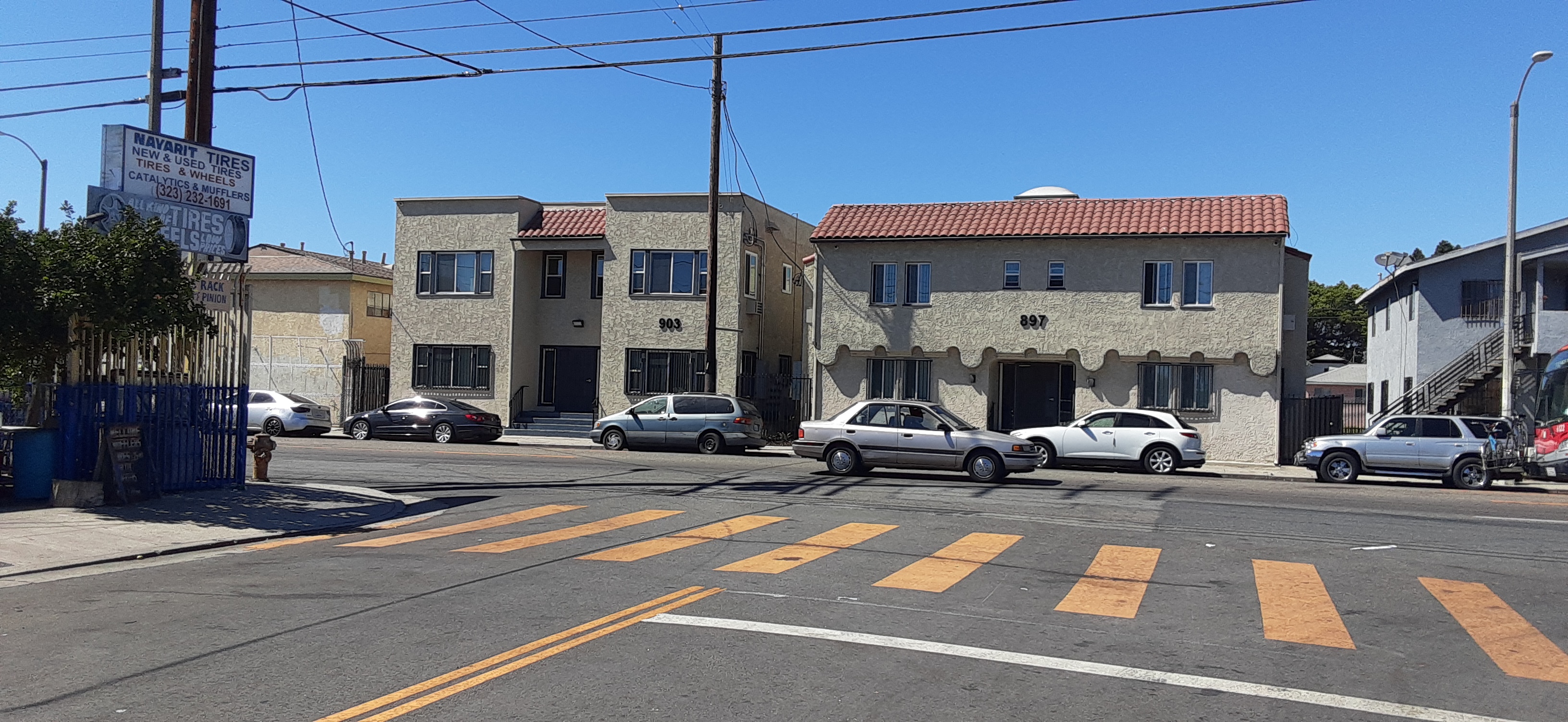 Front view of two beige 2 story buildings next to each other with a black steel gate in the middle, multiple windows on both building, the bottom ones gated, front doors with black steel handrails, five cars parked in front of the buildings, one car drivi