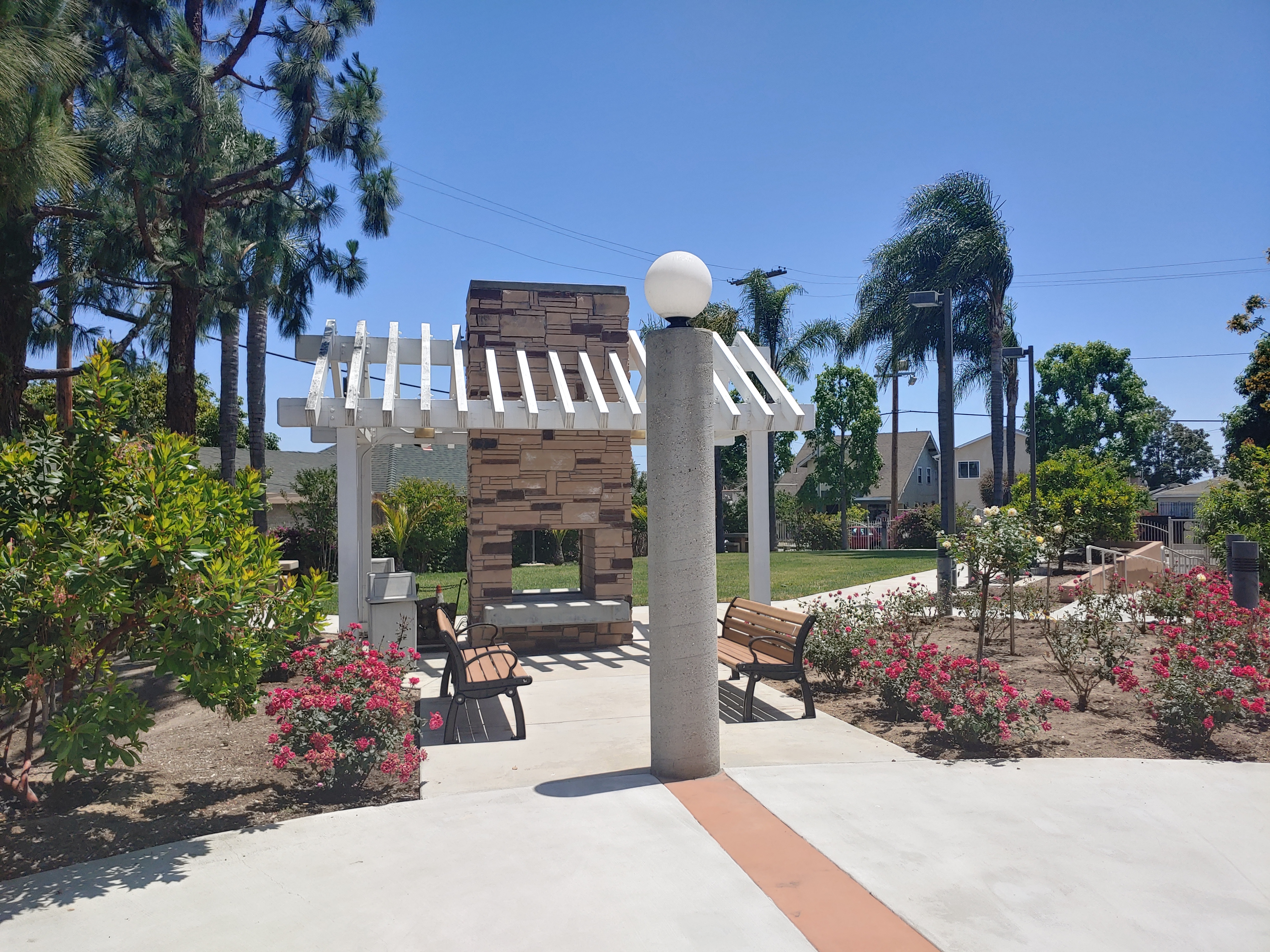 View of rose garden, two benches, white pergola, and built in outdoor fireplace with a grass area in the background.