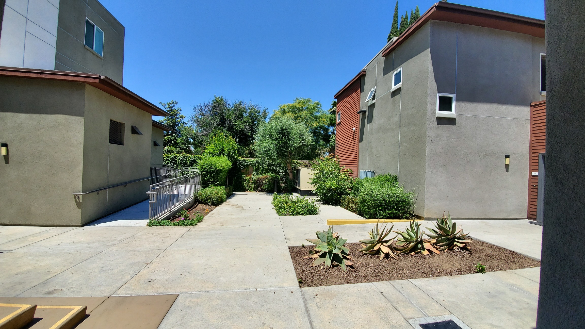 Different view of courtyard. There are various bushes, plants and trees. There is a ramp to access some units, and small steps to access another section of units.