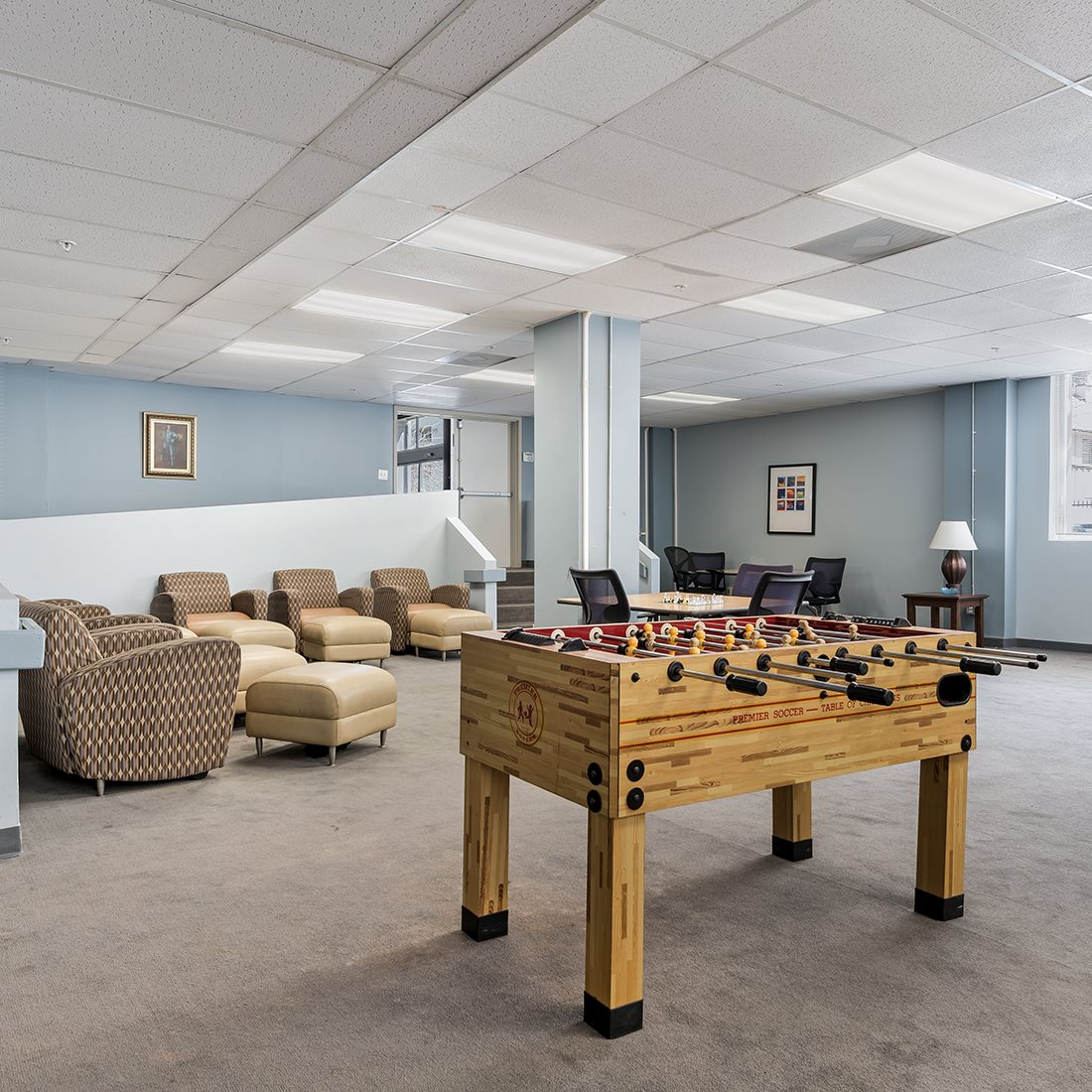 Community room with a foosball table, two round tables with chairs, and multiple sofa chairs. Room has stair and ramp access. Floor is carpeted.