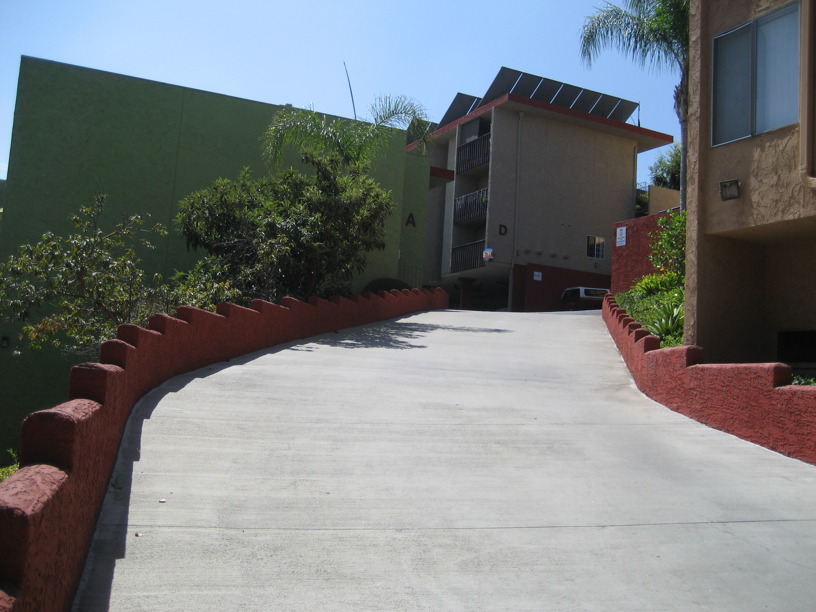 View of inside of the driveway. There is a small red wall on both sides and trees visible from behind the walls. Driveway leads to an outdoor parking lot.