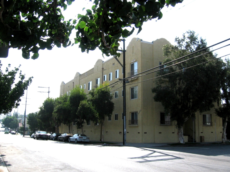 Three story yellow building located on corner. Trees surround the outside providing significant shade with cars parked on street. Front entrance has stair access with grab bars.