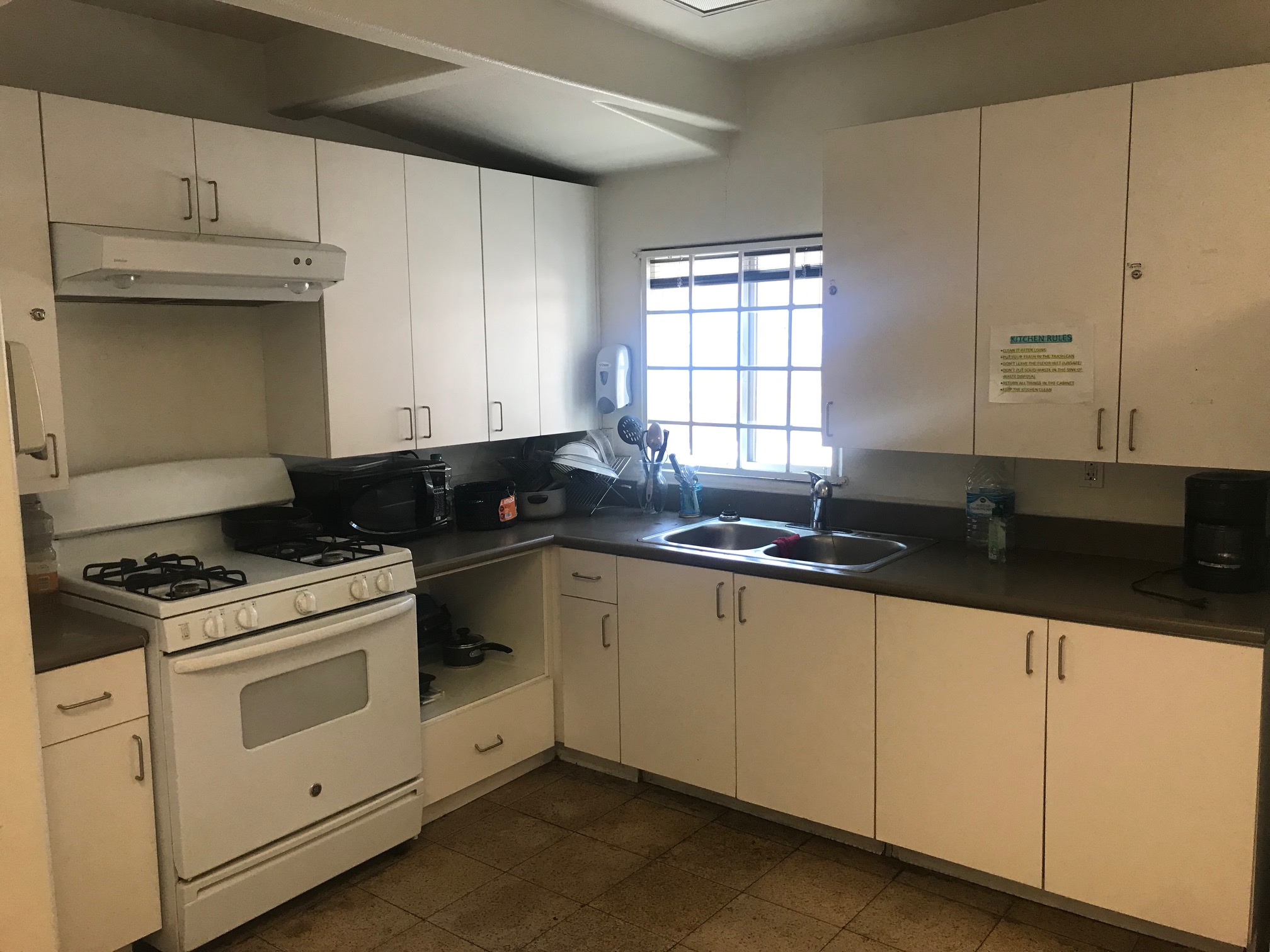 Left side view of a kitchen, white pantry, white stove with oven and under cabinet range hood, black microwave, pots, dishes, gallon of water, cooffe maker, soap dispenser, white window.