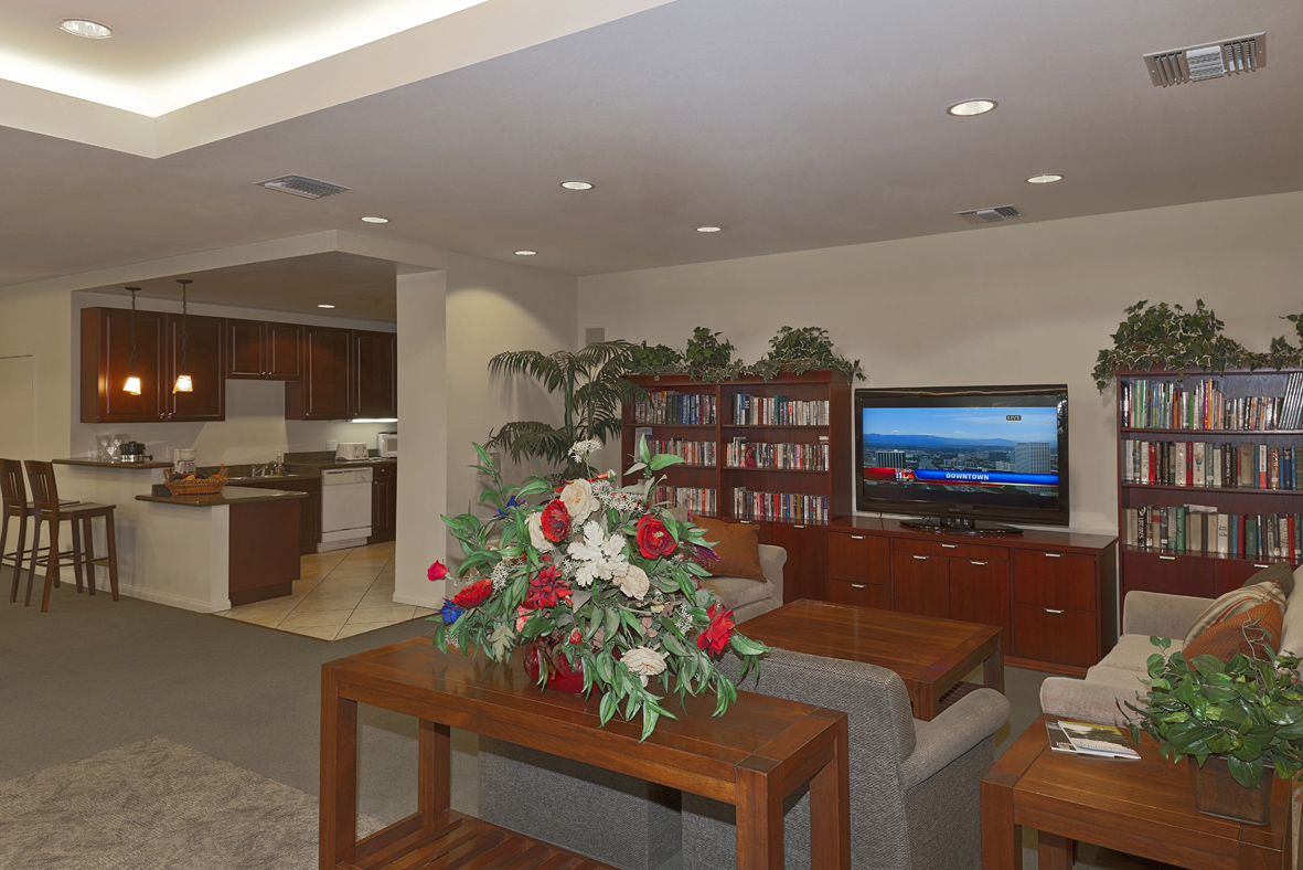 Community room with a kitchen area and lounging area. Lounging area has sofas, a coffee table, flat screen TV, and a large bookshelf. There is also a couple of plants on top of side tables. Kitchen area has a counter with two high wooden chairs, From this