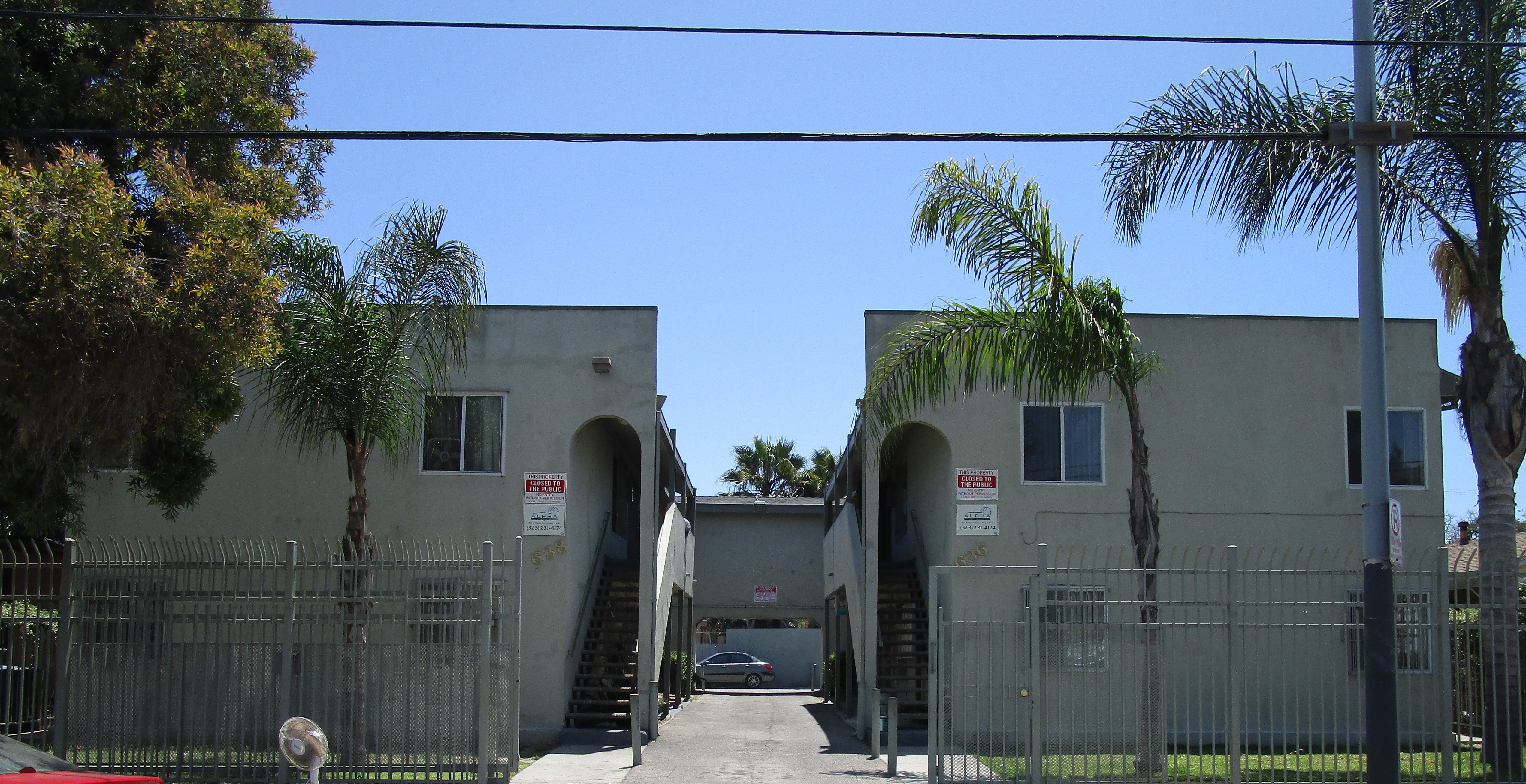 Front view of two apartment buildings next to each other, four windows on each building, two story, stairs up on both buildings, open gate, grass in front of the units, multiple trees, far back a parked car in the parking lot, no public parking and manage