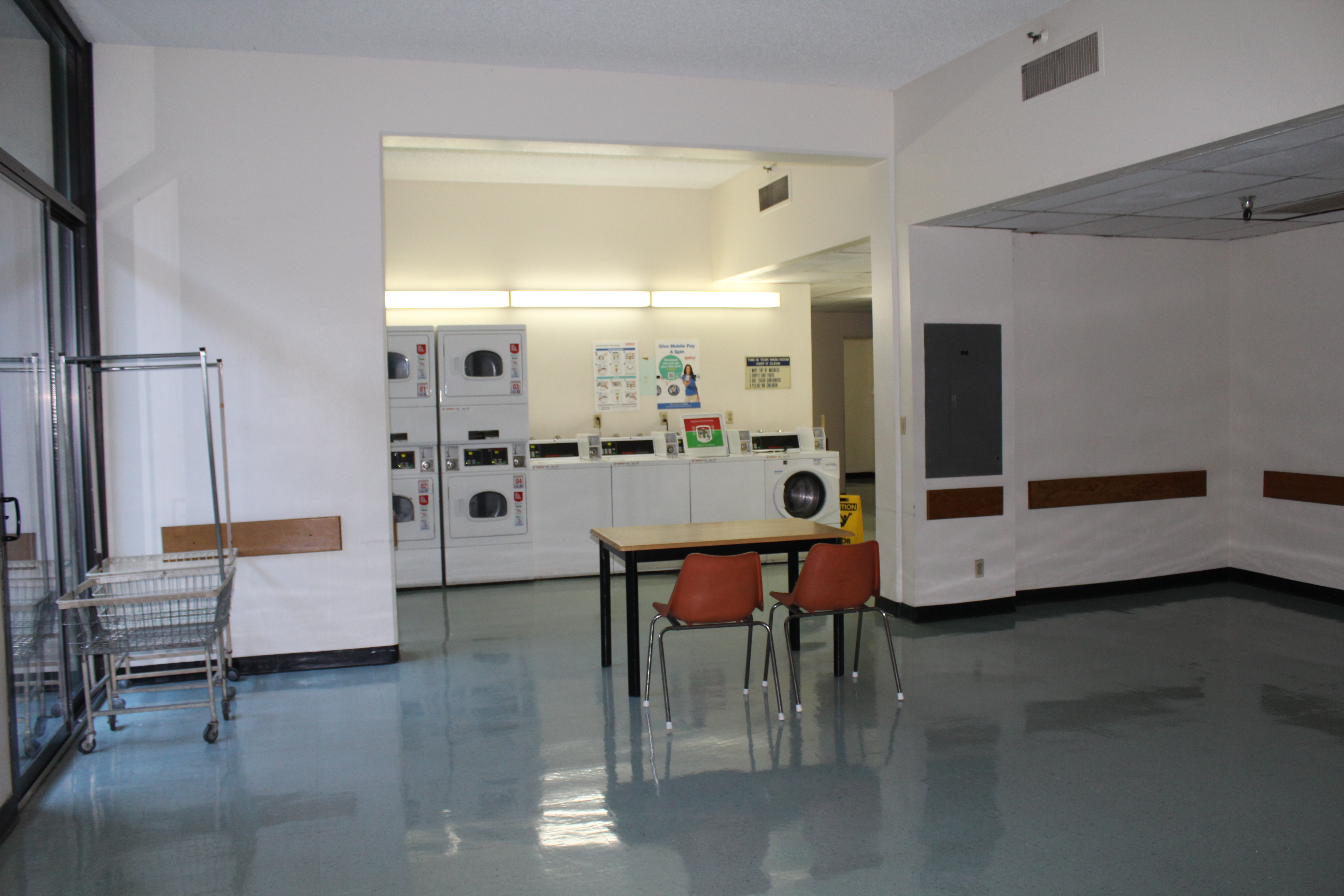 Interior view of a laundry room for Angelus Plaza 1. Large comparmental area with washers and dryers, wire rolling basket and a table with two chairs. White walls with a brown boarder.